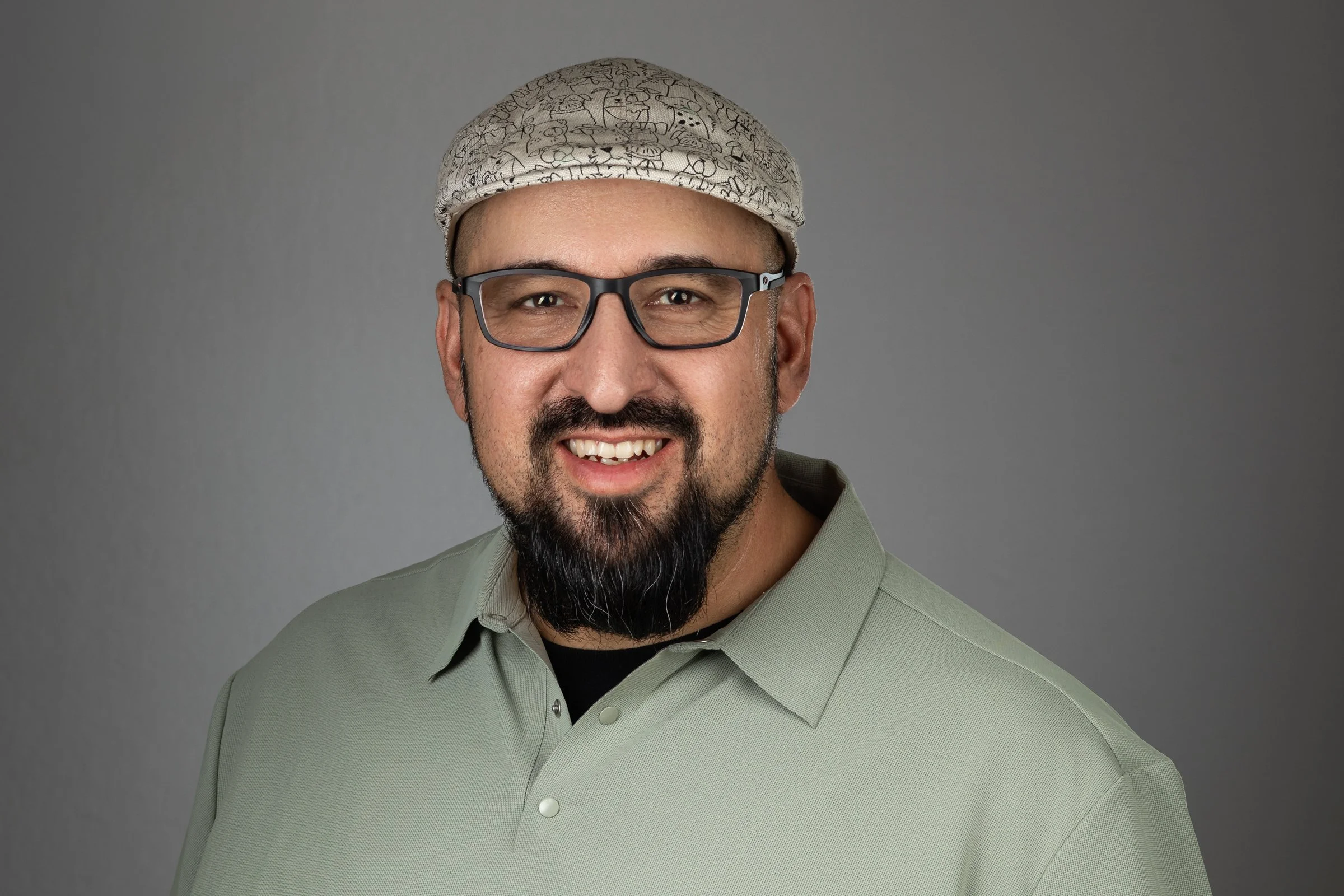 A man with glasses, a beard, and a mustache wearing a light green polo shirt and a patterned cap, smiling against a gray background.