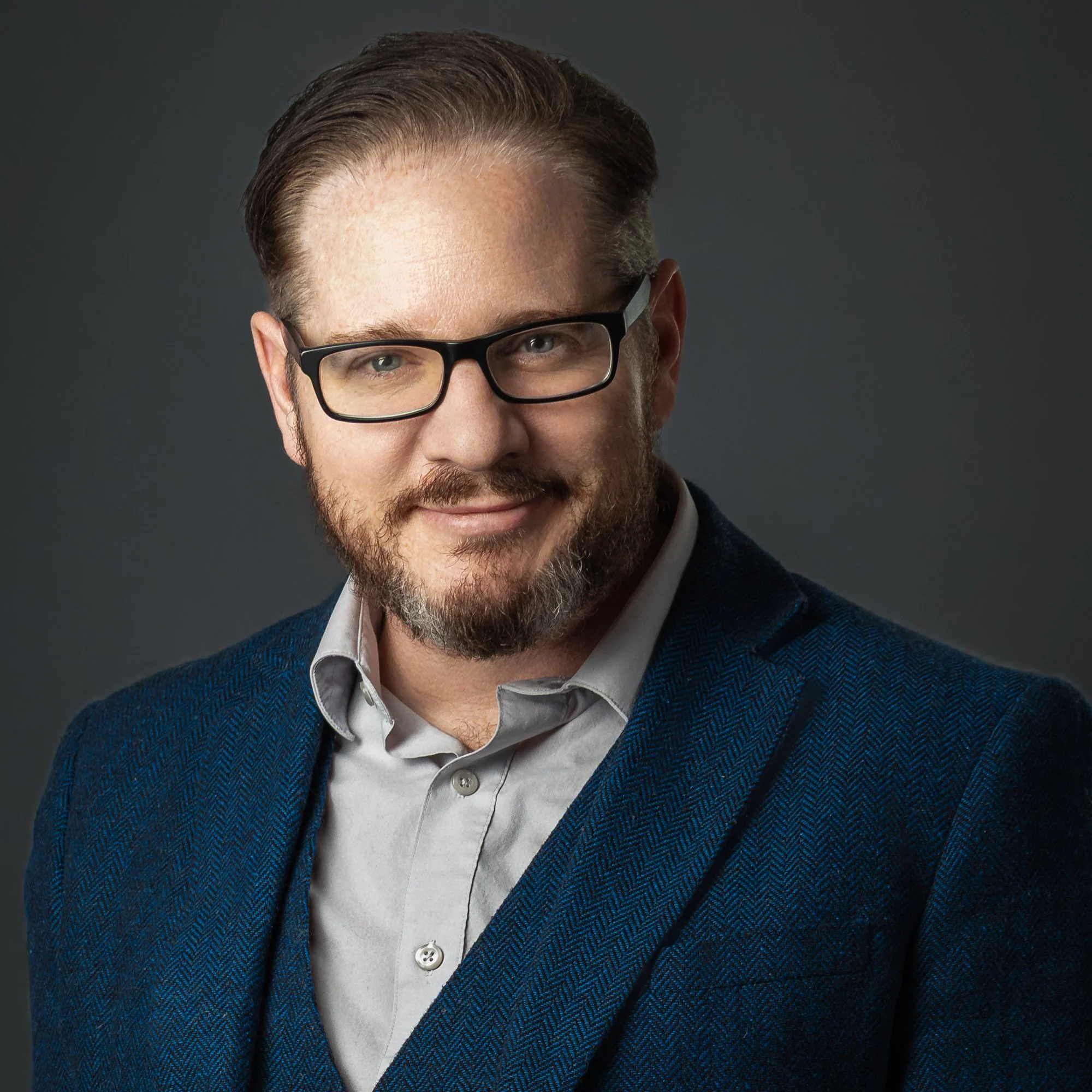 Close-up of a man with glasses and facial hair wearing a white shirt and a dark blue blazer against a dark background.