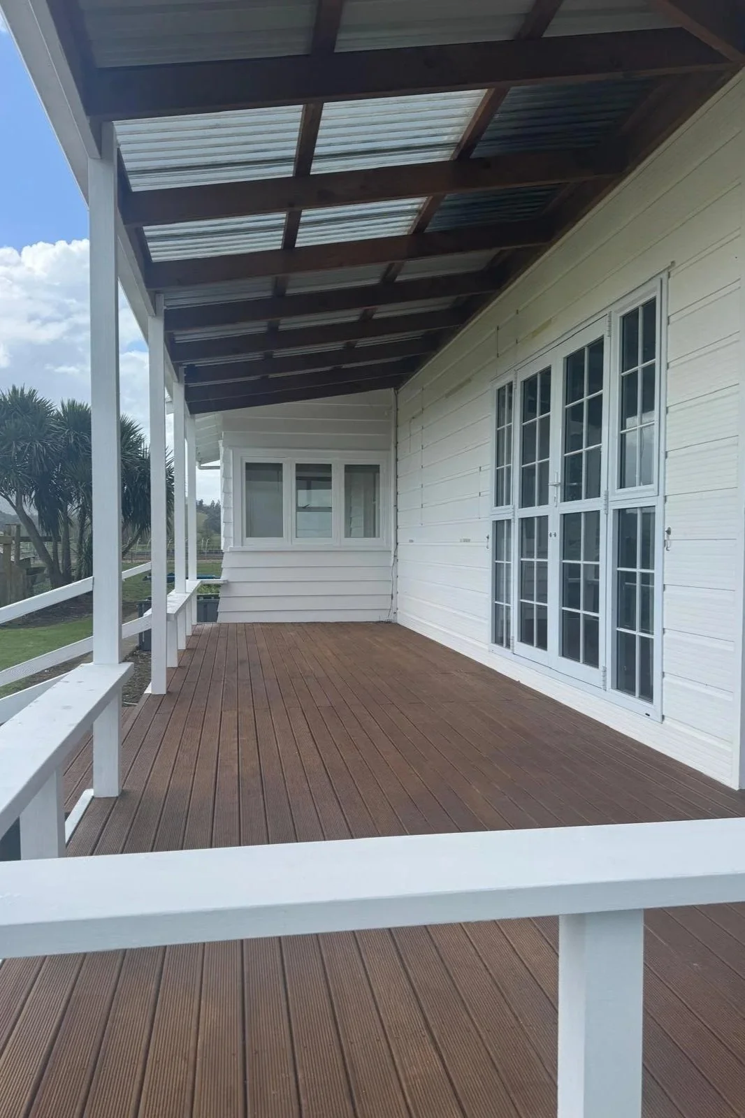 A wooden balcony with white support beams, a white railing, and a paper roof, attached to a house with white siding and several large windows.