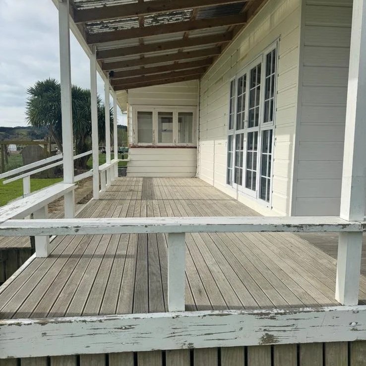 View of a weathered wooden porch with a railing, attached to a cream-colored house with large windows and sliding glass doors.