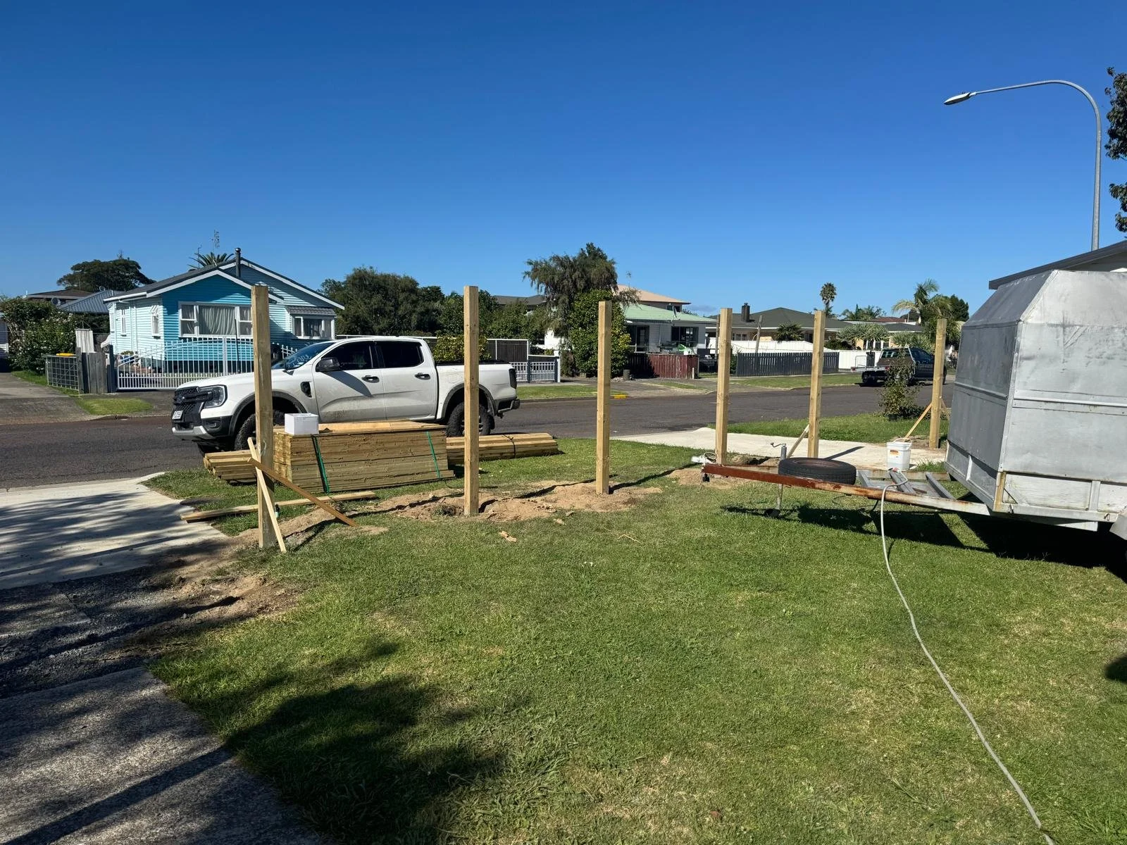 Residential front yard under construction with wooden posts for a fence, a trailer with building materials, and a white pickup truck facing the street. Houses and a street are visible in the background under a clear blue sky.