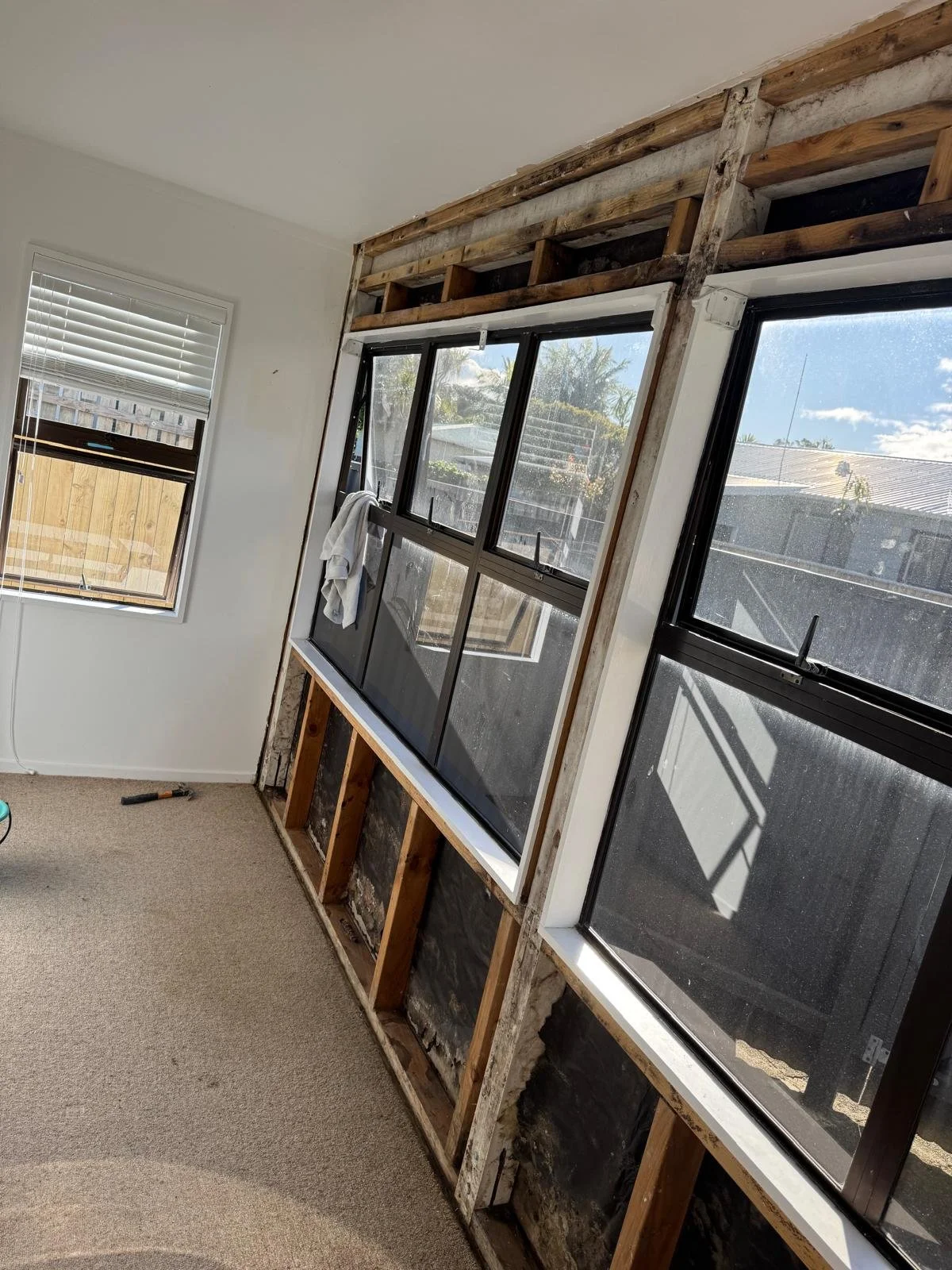 Room undergoing window remodeling showing some window frames and partially removed wall siding, with a towel hanging on one window and construction tools on the floor.