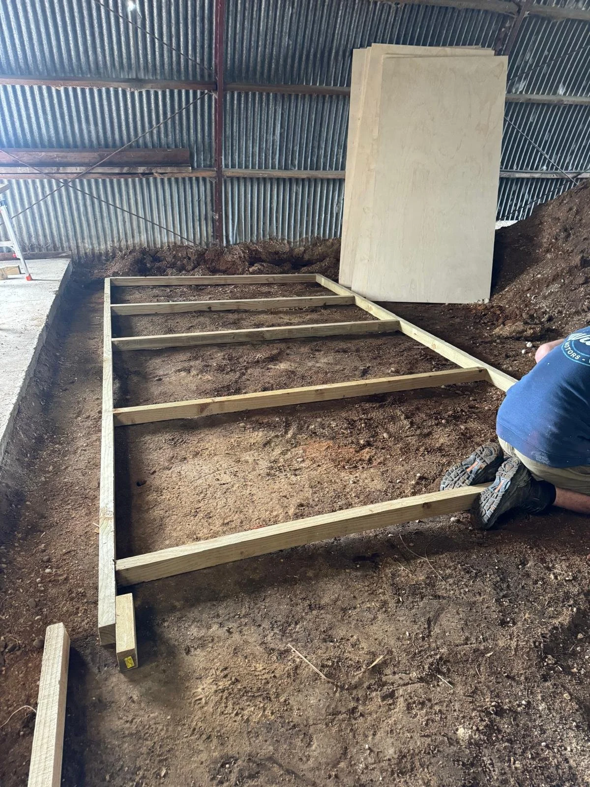 A person kneeling on the ground constructing a wooden frame on a dirt surface inside a building with corrugated metal walls and a partially visible ladder.