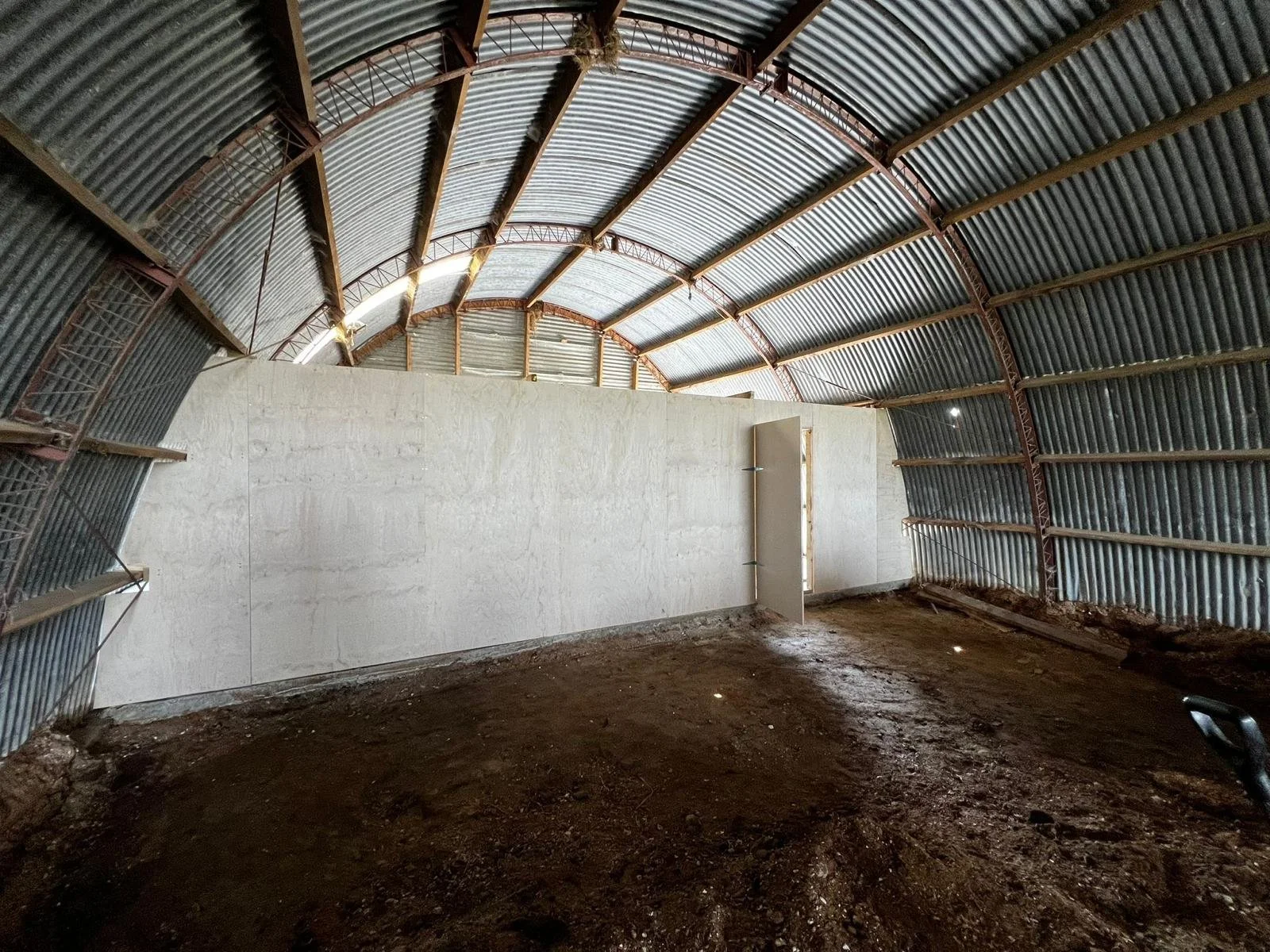 Inside a partially constructed metal building with curved corrugated metal walls and roof, a white partition wall is visible with an open door, and the ground is uneven dirt.