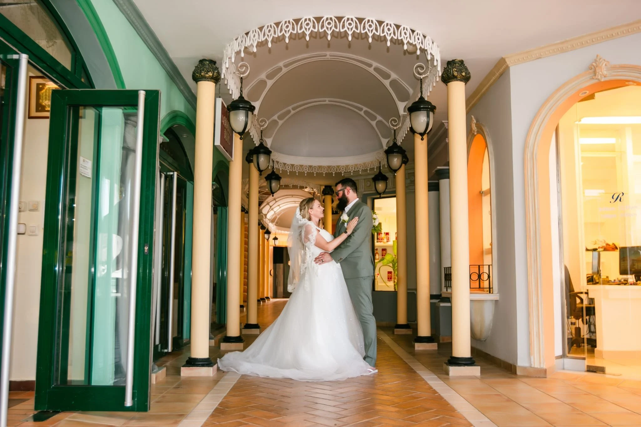 Bride and groom in wedding attire standing close inside a decorative hallway with columns and hanging lanterns, gazing at each other.