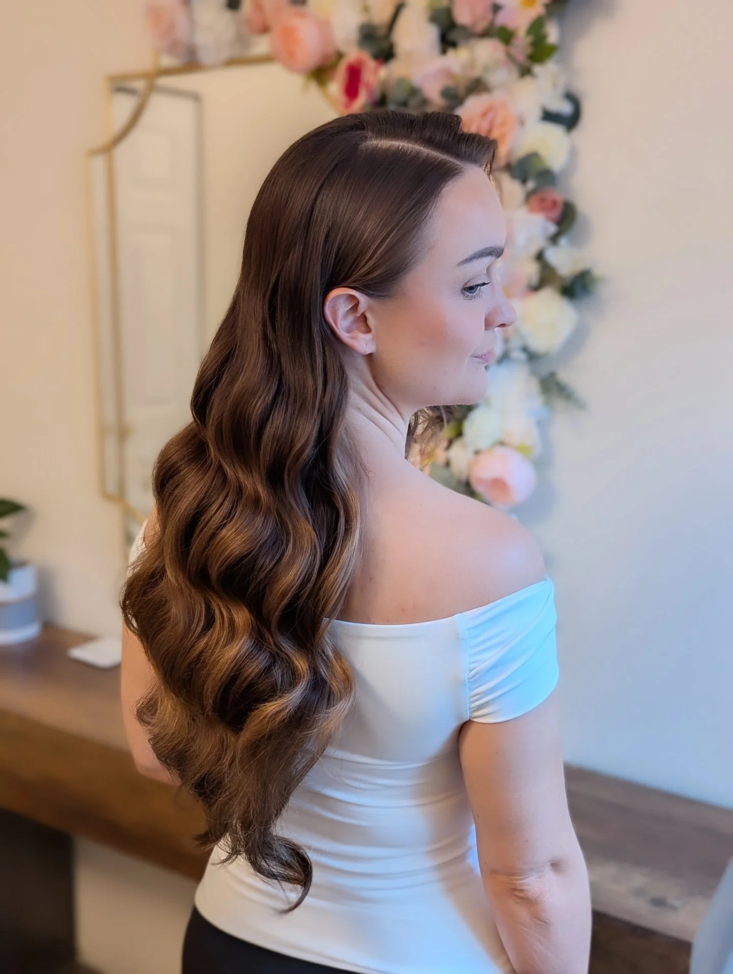 A woman with long, wavy brown hair is looking to her right, standing indoors in front of a flowered wall. She has a smooth off-the-shoulder white top.