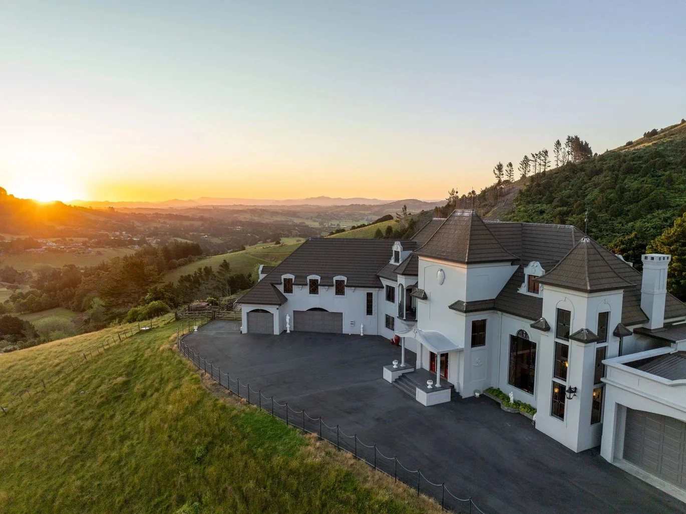 A large white mansion with a dark gray roof on a hillside during sunset, surrounded by green fields and trees.