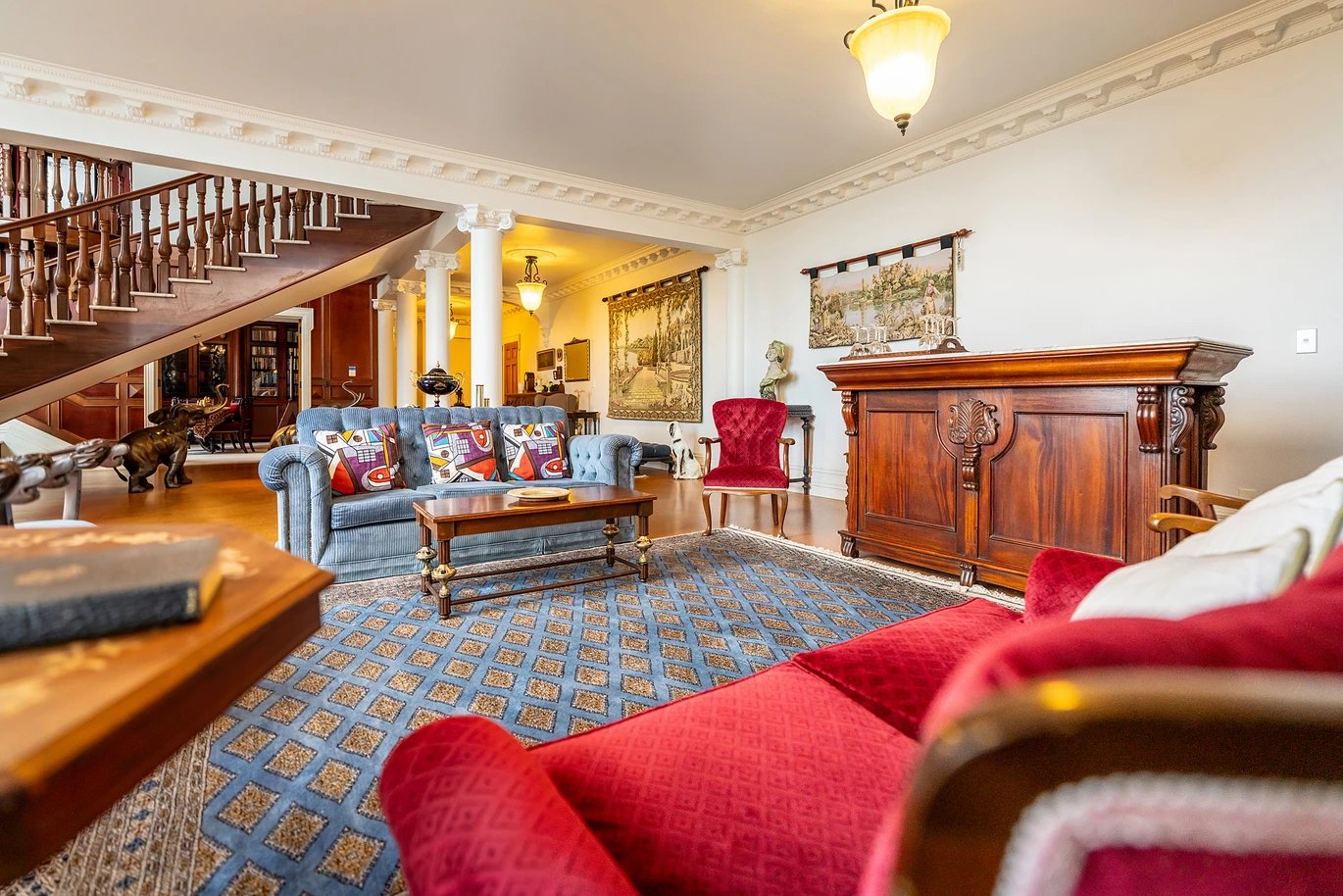 A spacious living room with a blue tufted sofa, red armchair, wooden coffee table, and an ornate wooden sideboard. Decor includes wall hangings, a stone sculpture, and decorative items. Hardwood floors and a patterned blue and gold rug are present, w