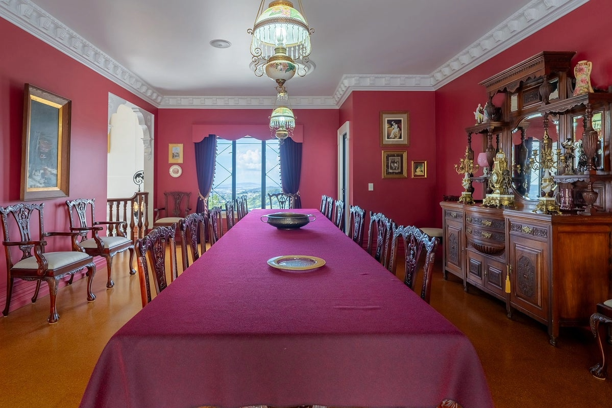 A dining room with red walls, a long table covered with a red tablecloth, and ornate wooden chairs. There are decorative items on a wooden sideboard and framed pictures on the walls. A large window shows a view of the outdoors, and hanging light fixt