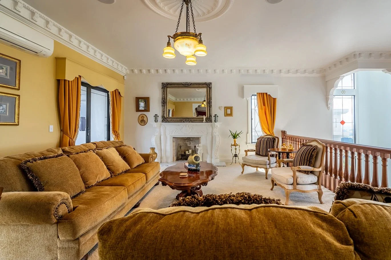 Living room with beige and white walls, vintage furniture, a marble fireplace with a mirror above, gold curtains, and a chandelier.