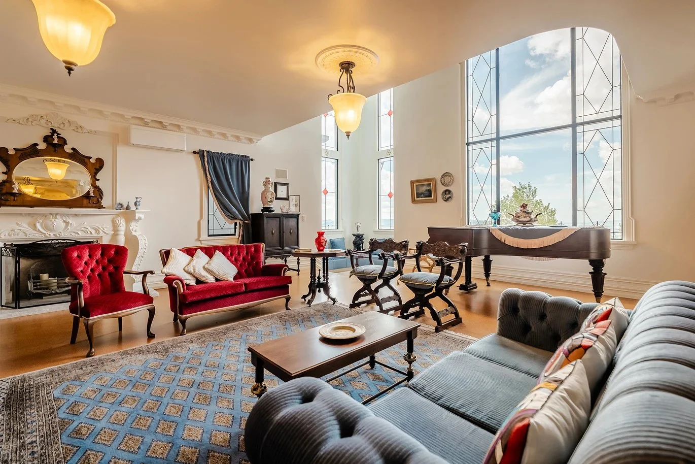 A spacious living room with vintage furniture, including a gray sofa, a red velvet loveseat, and matching armchair, a wooden coffee table, and a grand piano near large windows with a scenic view. Decor includes framed art, a marble fireplace, and cei