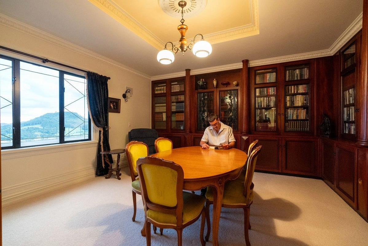 Man sitting at a wooden conference table in a library or study room, with bookshelves on the wall and a window showing a view of a mountain outside.