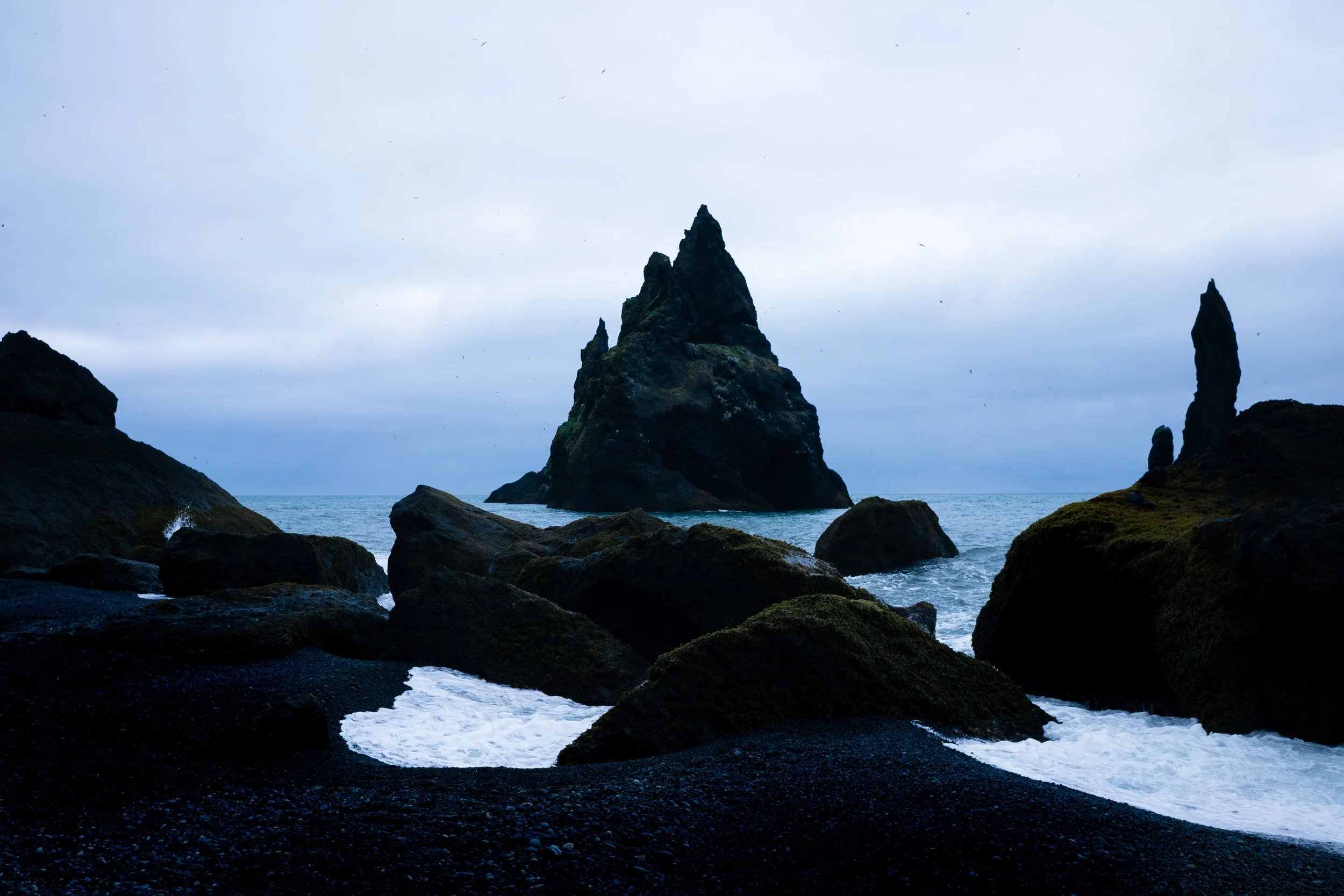 A rocky coastline with black sand and large moss-covered rocks, featuring prominent sea stacks in the background under a cloudy sky.