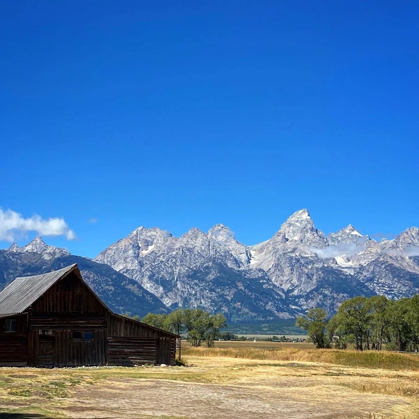 A rustic wooden barn in front of a mountain range with snow-capped peaks and a clear blue sky.