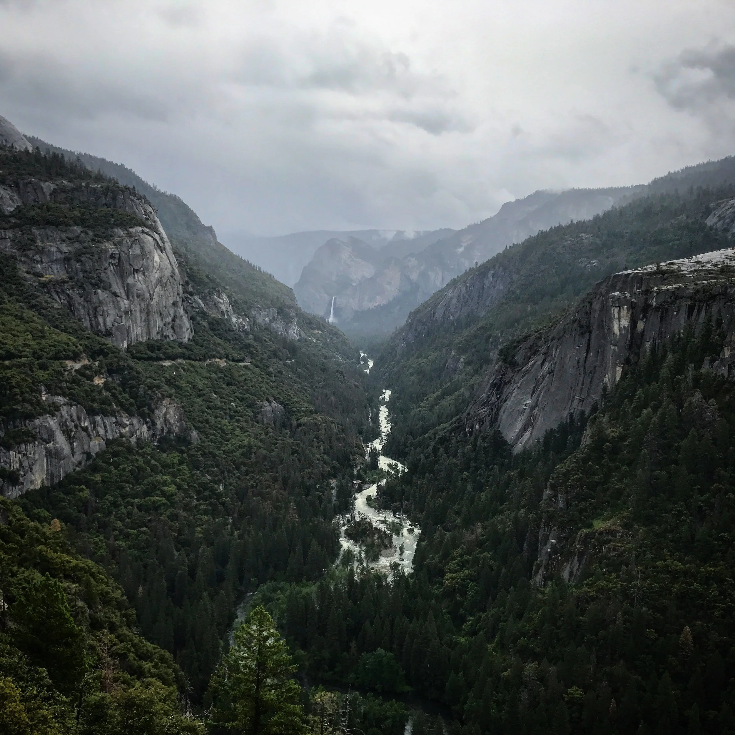 A view of a deep valley with a river running through it, flanked by steep, forested mountains under a cloudy sky.