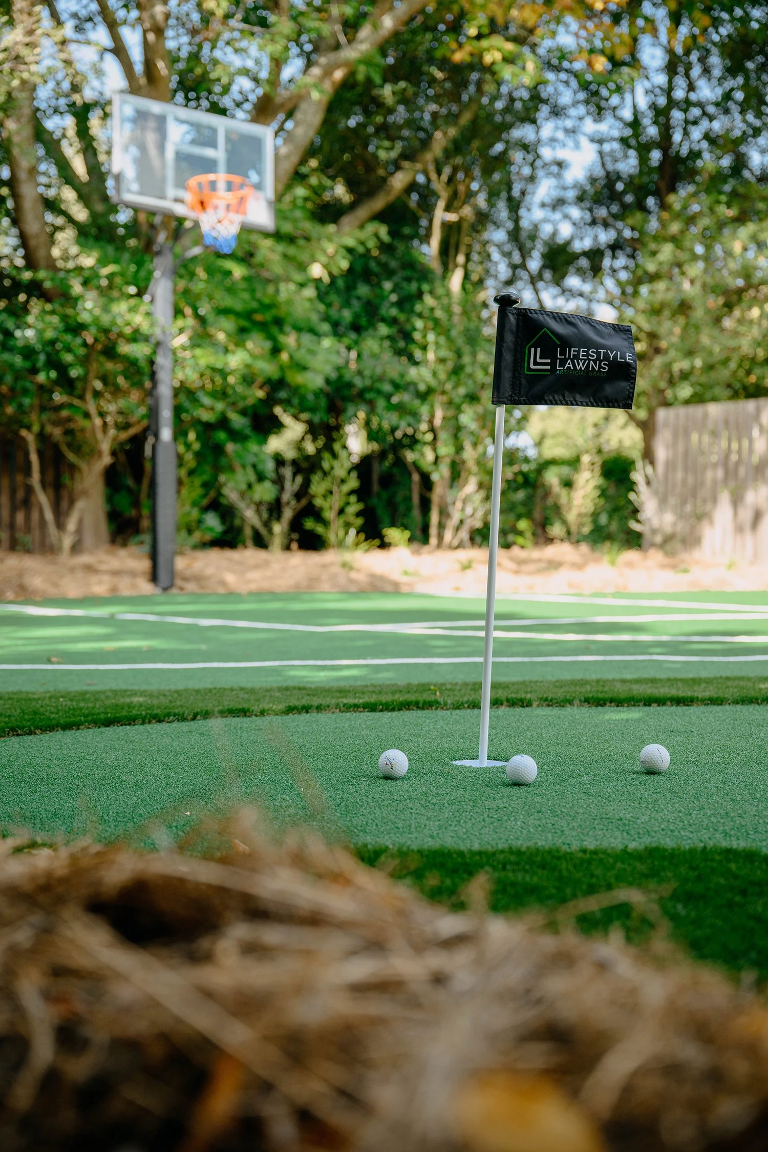Backyard golf putting green with golf balls near the hole and a flag marked 'Lifestyle Lawns' in a backyard with a basketball hoop and trees in the background.