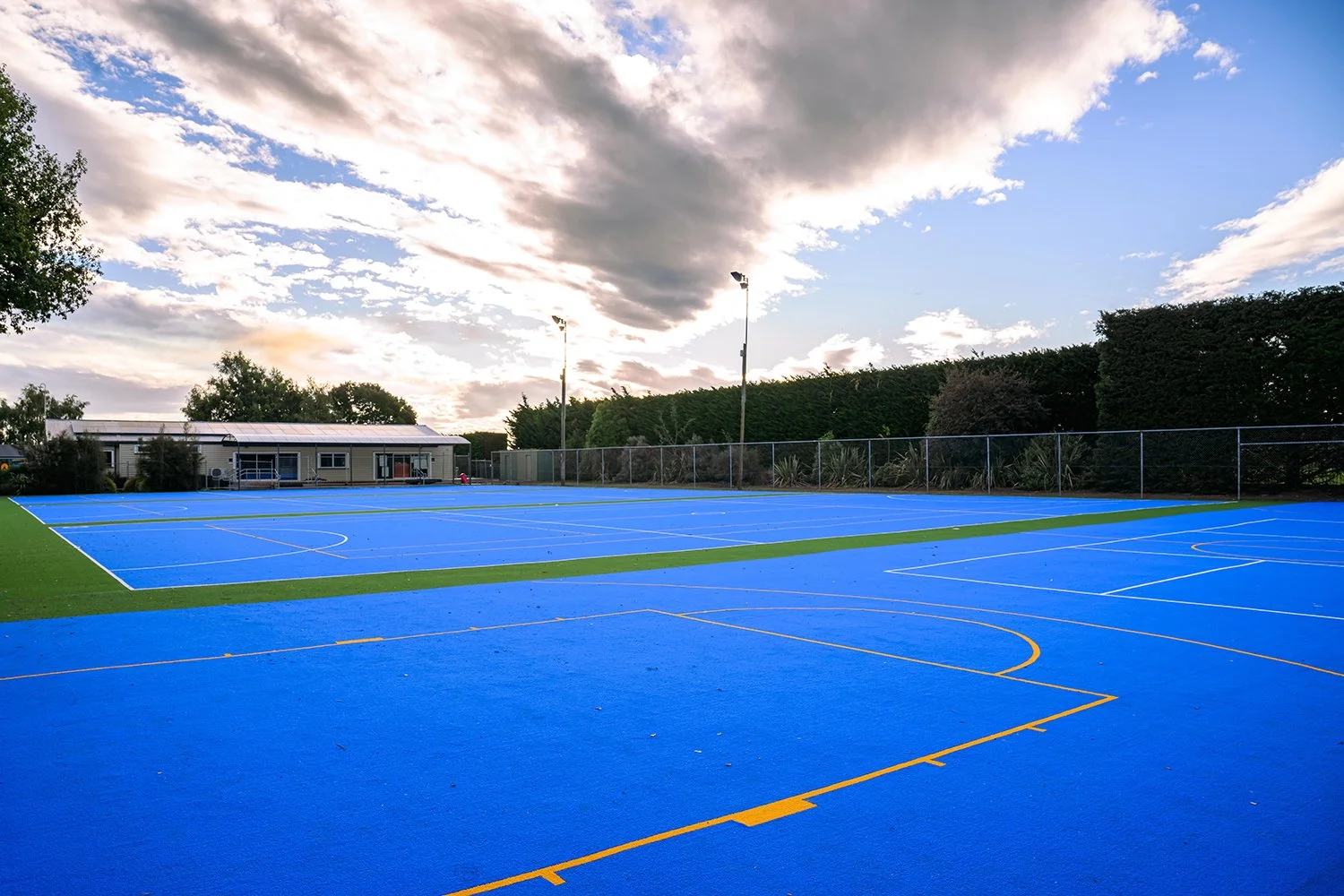 A blue, school multi sport court with white and yellow lines, surrounded by greenery and a fence. There is a classroom in the background and a cloudy sky above.