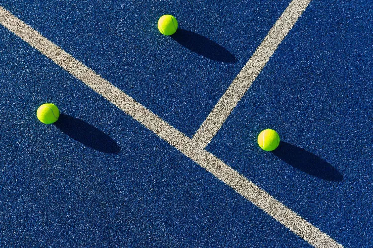 Three yellow tennis balls on a blue tennis court with white boundary lines, casting shadows.