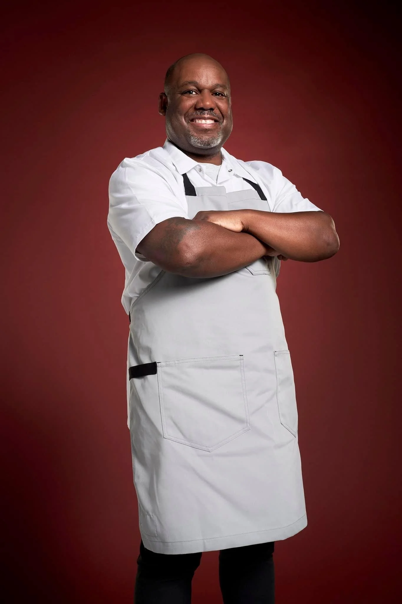 A man smiling with arms crossed, wearing a white apron over a white shirt, against a dark red background.