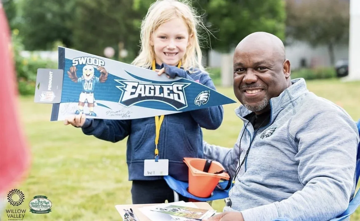 A young girl and a man outdoors, holding a Philadelphia Eagles football pennant. The girl is smiling, wearing a blue jacket and has a lanyard, and the man is sitting in a wheelchair, wearing a gray jacket with Eagles logo, smiling at the camera.