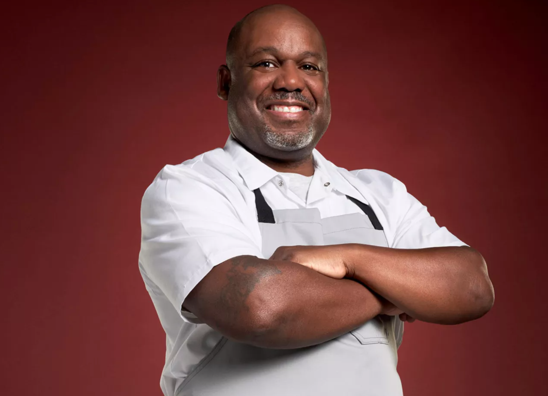 A confident chef Mark McMillian with arms crossed, wearing a white chef's jacket, standing against a dark red background.