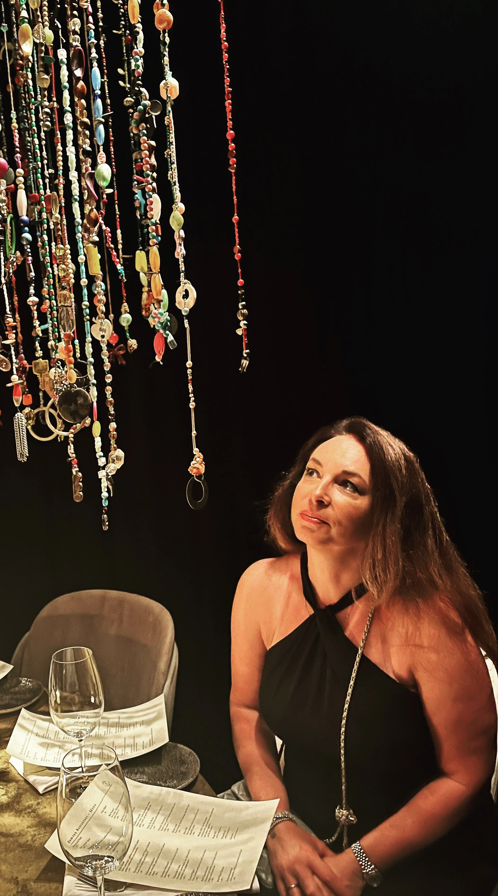A woman with long brown hair in a black dress sitting at a table with wine glasses and menus, looking up at a display of various colorful necklaces hanging from the ceiling against a black background.