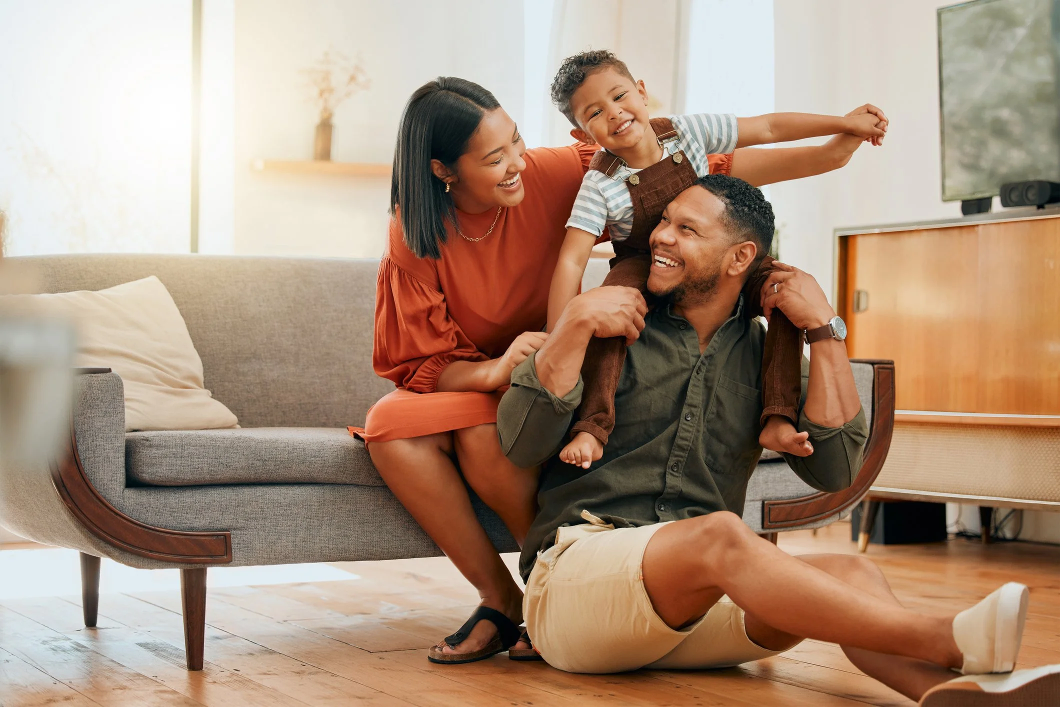 A family of three playing and smiling in their living room, with the mother sitting on a couch, the father sitting on the floor, and the young son balancing on the father's shoulders.
