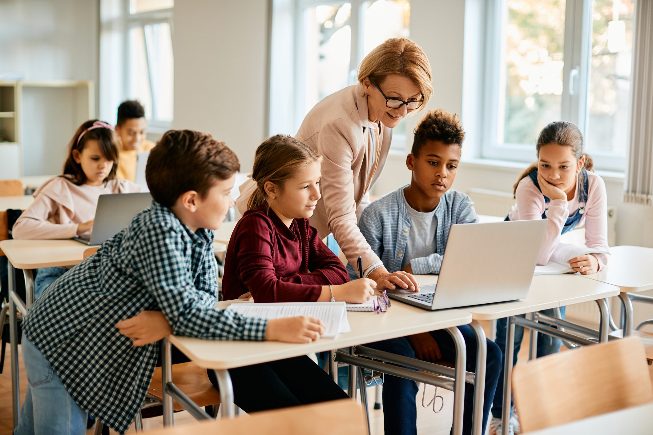 A teacher assisting students in a classroom with a laptop. Six children are seated at desks, some taking notes or working on their laptops, with a bright classroom and large windows in the background.