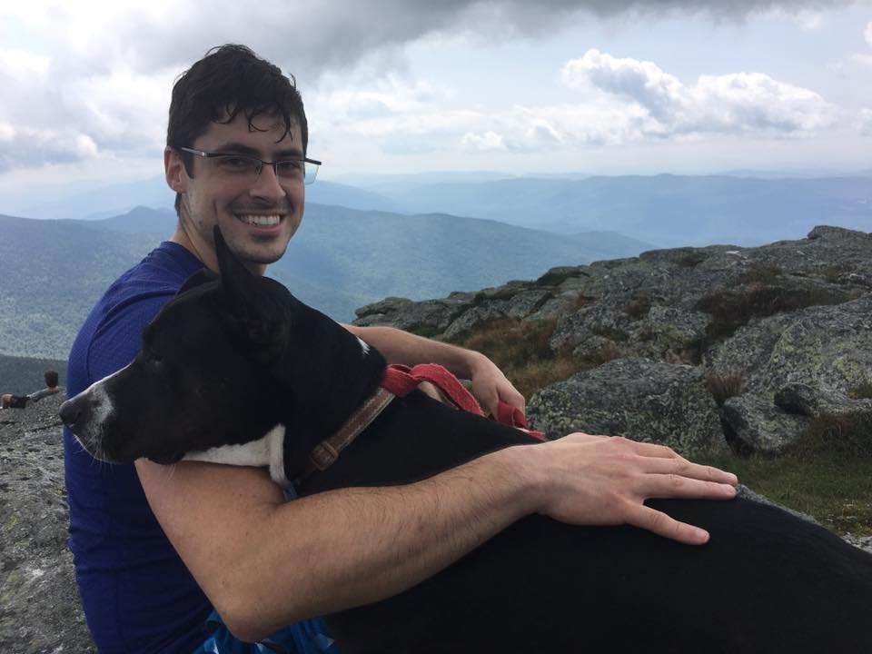 A smiling man in glasses and a blue shirt sitting on a rocky mountain top, holding a black dog with white markings around the nose and neck, surrounded by mountains and cloudy sky in the background.