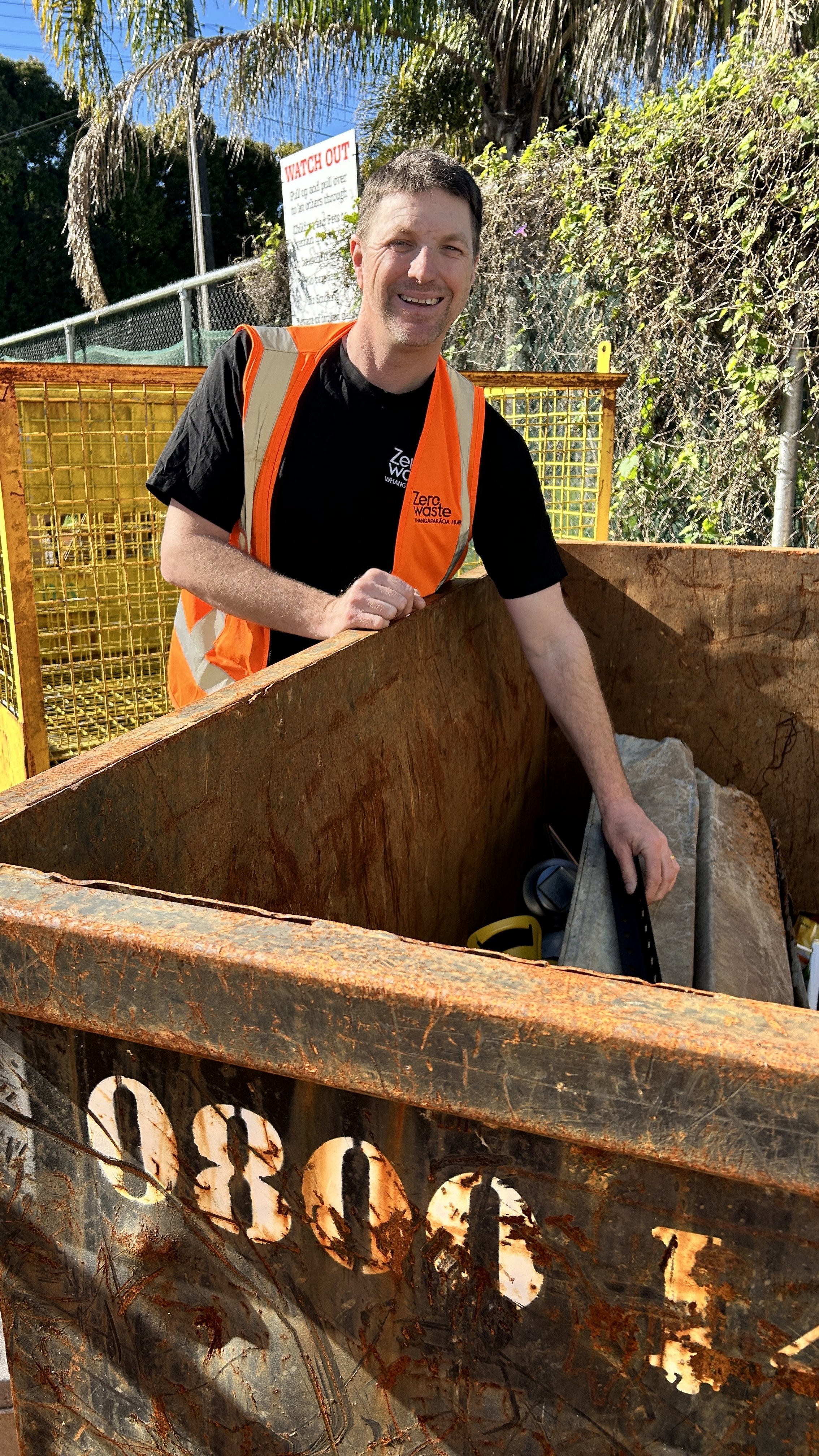 A man wearing a black shirt and an orange high-visibility vest standing next to a rusty metal skip container outdoors. He is smiling and holding a pair of black clippers, with trees and a sign in the background.