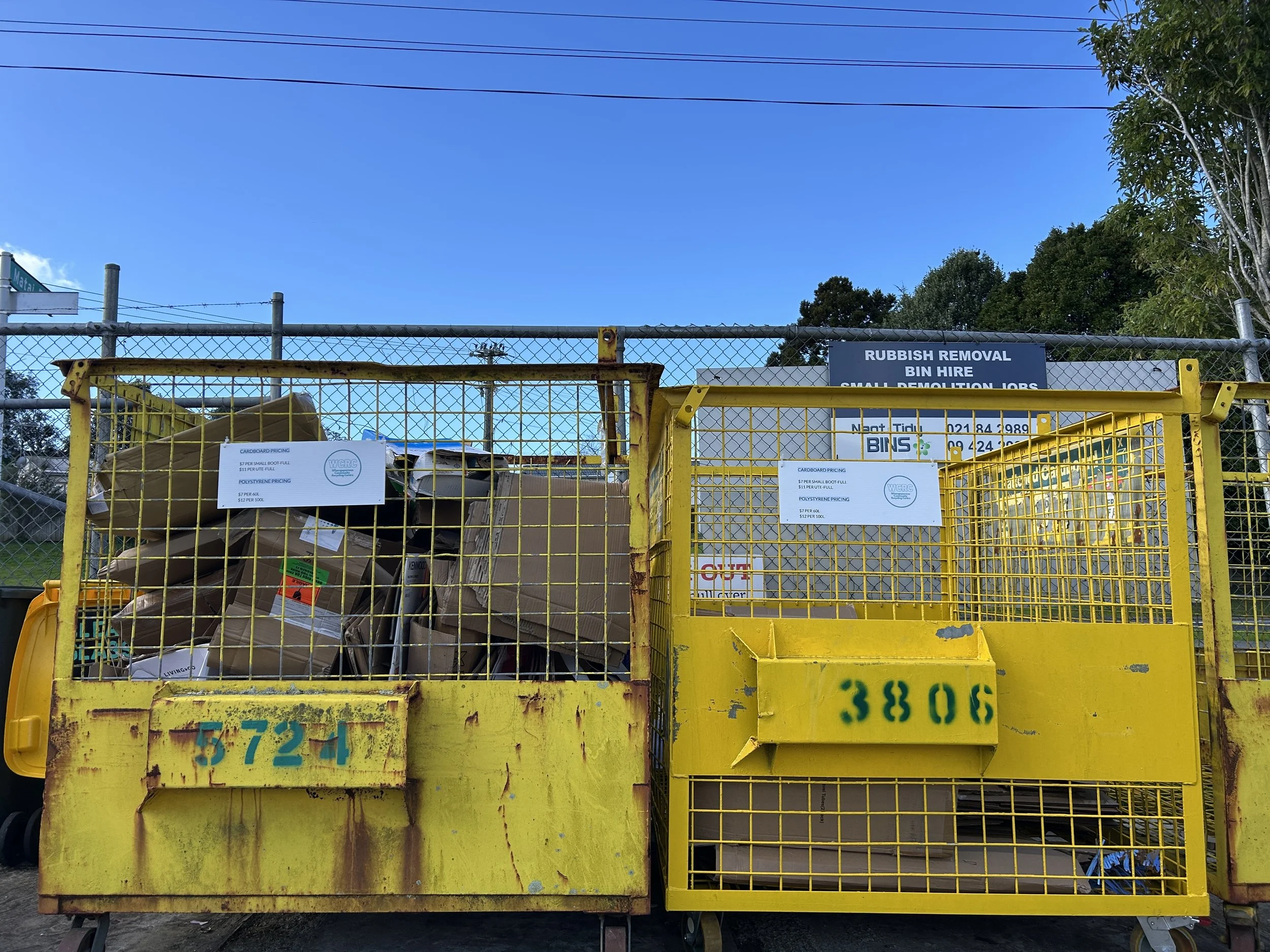 A yellow dumpster filled with cardboard and recycling materials, surrounded by a chain-link fence, with signs offering rubbish removal and bin hire services, set against a backdrop of trees and a clear blue sky.