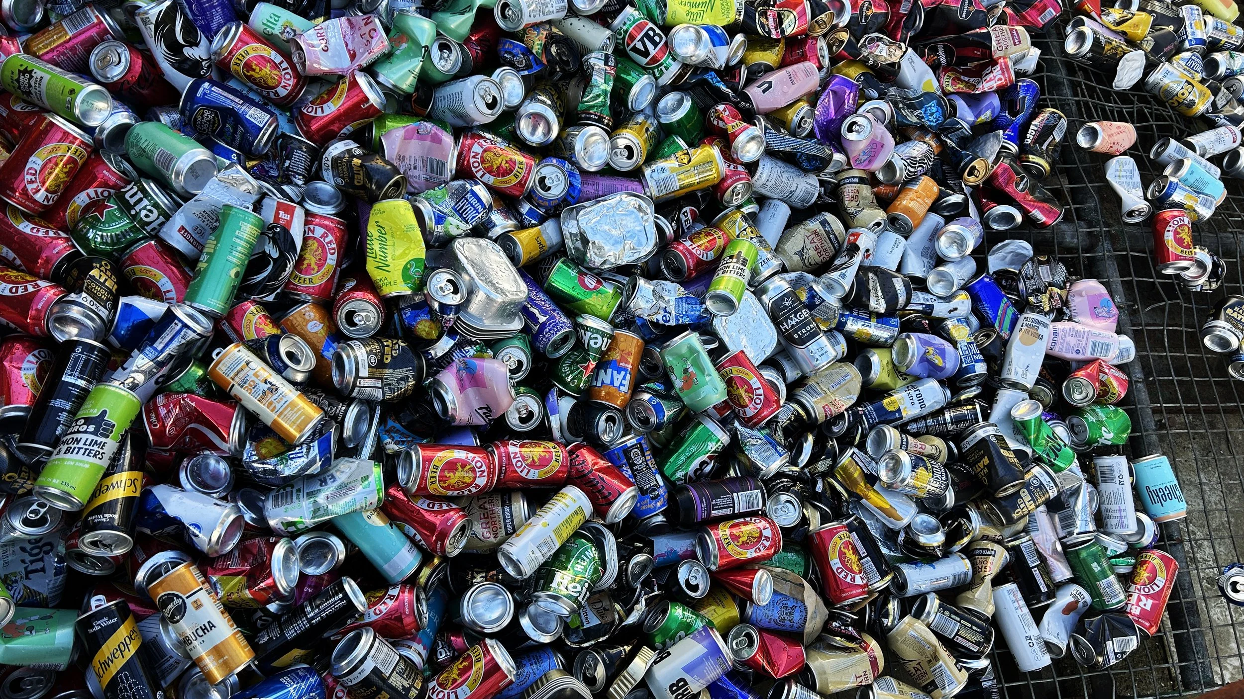 A large pile of crushed and intact aluminum cans, including brands such as Heineken, Red Bull, and Schweppes, on a metal grate surface, ready for recycling.