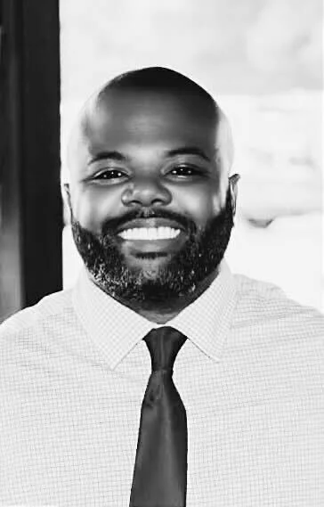 A smiling man with a beard in a collared shirt and tie, standing near a window.