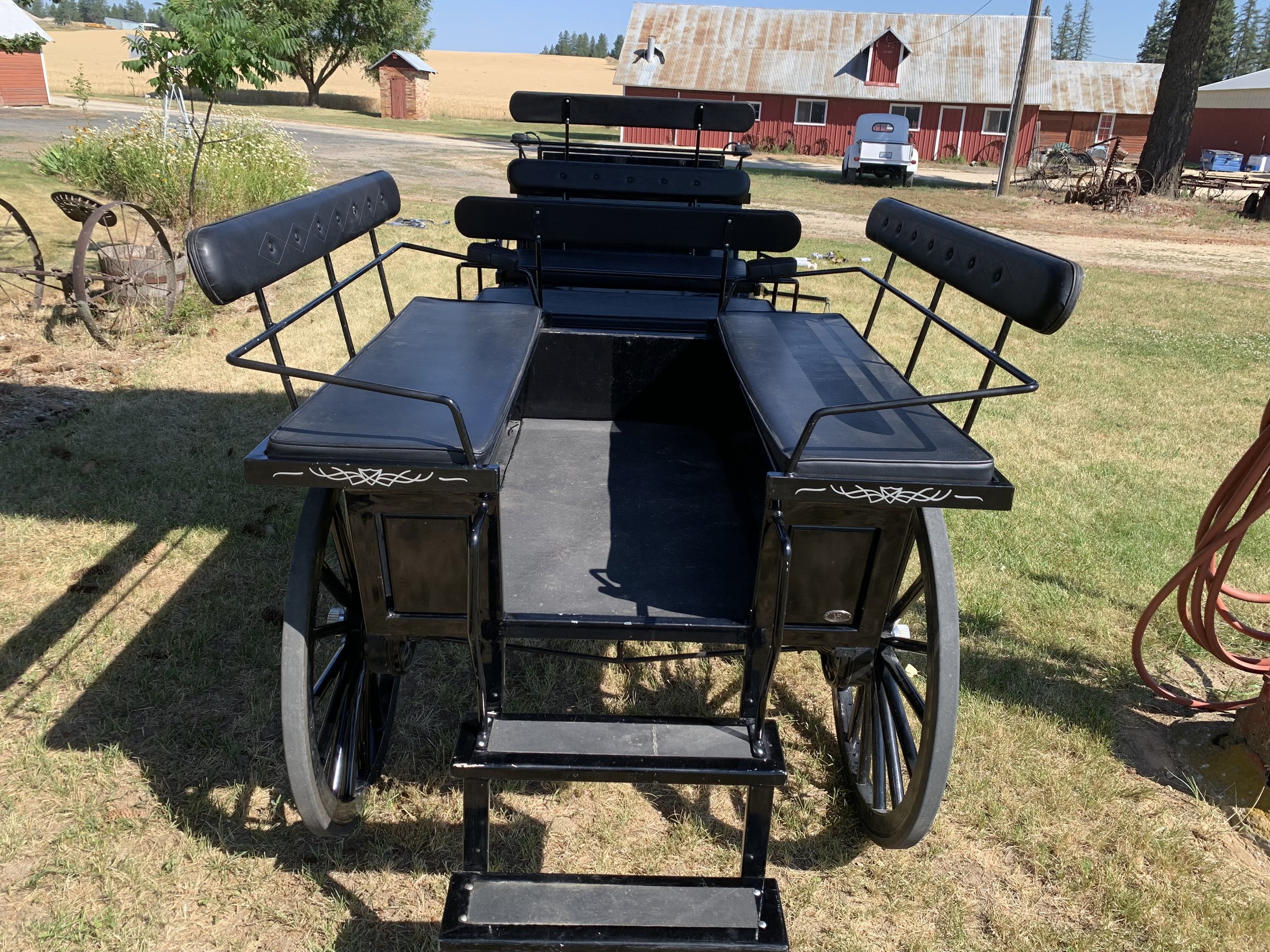 A vintage horse-drawn carriage with black leather upholstery and metal railings, parked on a grassy area outside a farm with red barn and countryside in the background.