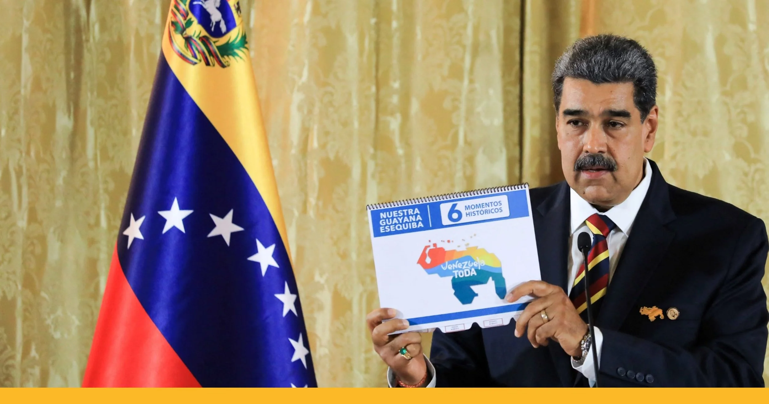 Hombre con bigote y cabello oscuro usando traje oscuro, camisa blanca y corbata con rayas rojas, negras y amarillas, sosteniendo una cartulina con un logo que dice 'Venezuela Toda' y un mapa del país. Detrás, bandera de Venezuela con franjas amarillas, azules y rojas, y estrellas blancas en un círculo. Fondo de cortinas doradas.