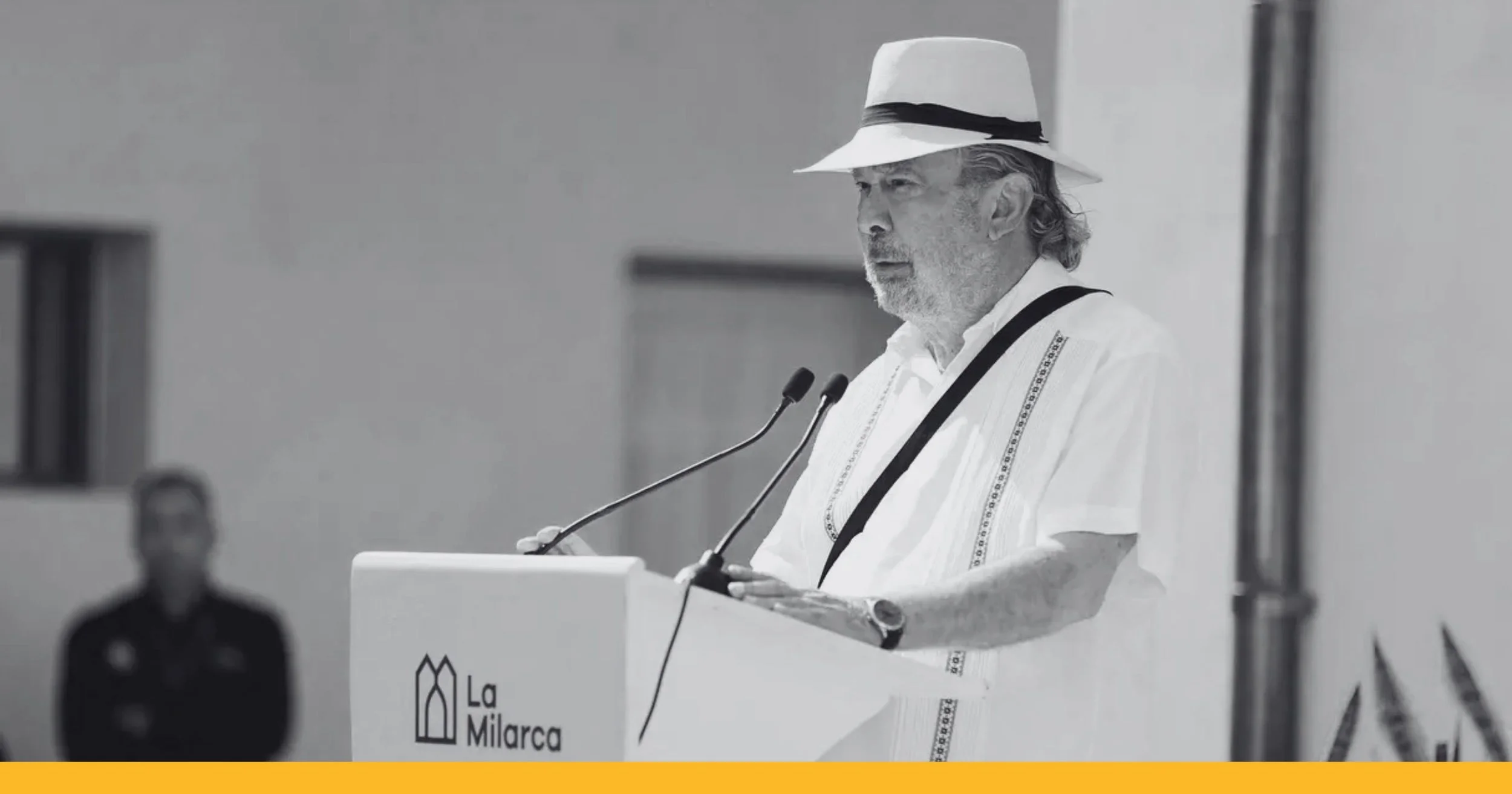 Hombre mayor con barba, gafas y sombrero blanco, hablando en un podio con el logotipo de La Milarca, en un evento público.