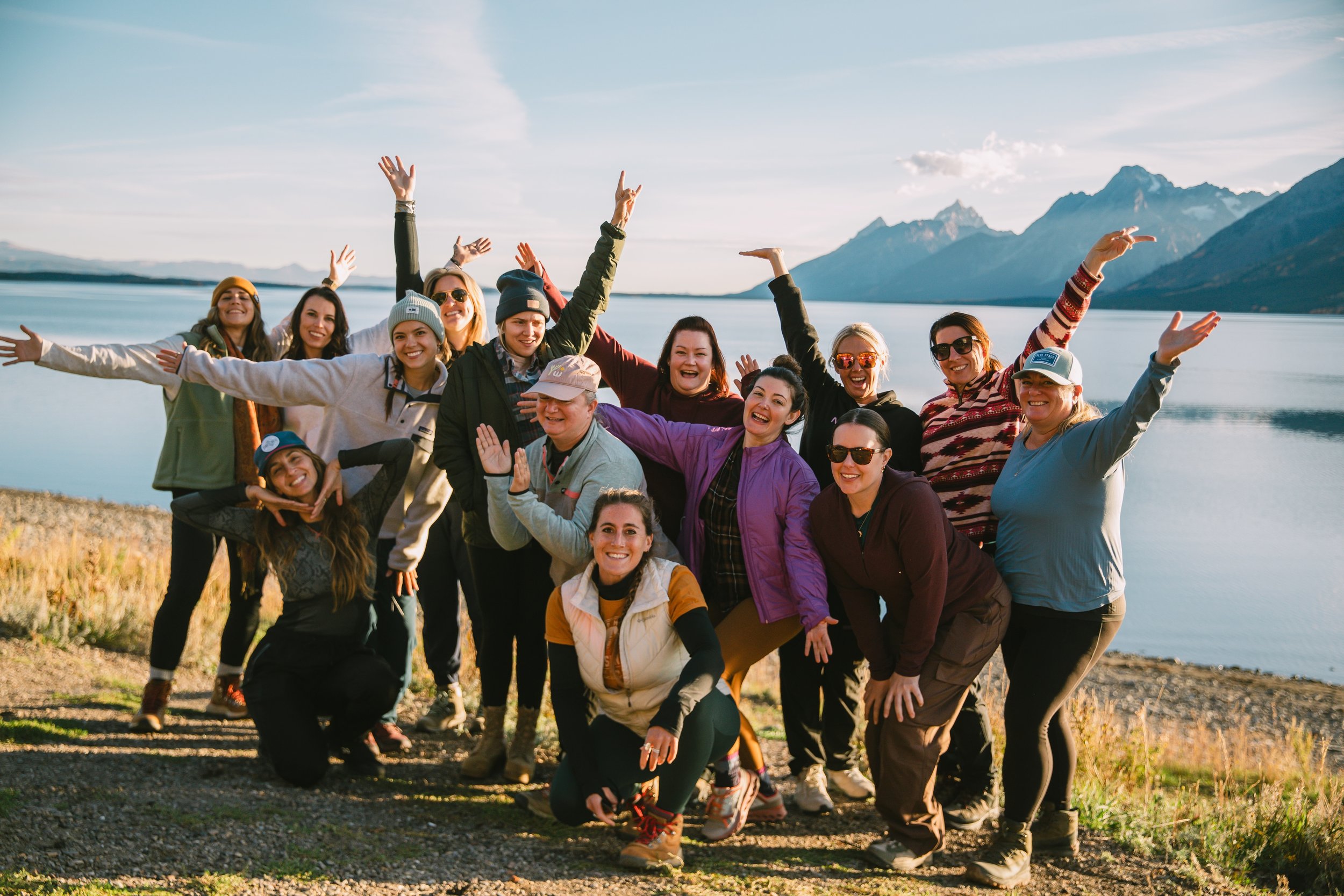 A group of women posing cheerfully outdoors near a lake, with mountains in the background.