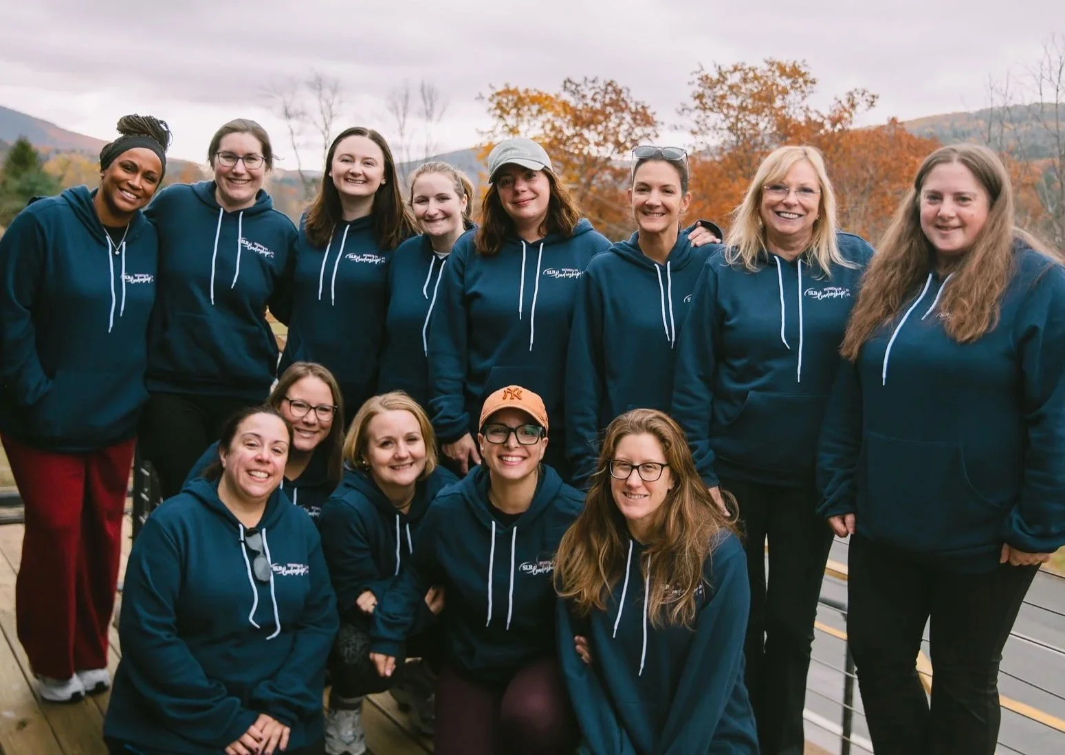 A group of 13 women wearing matching navy blue hoodies posing outdoors with autumn trees and mountains in the background.