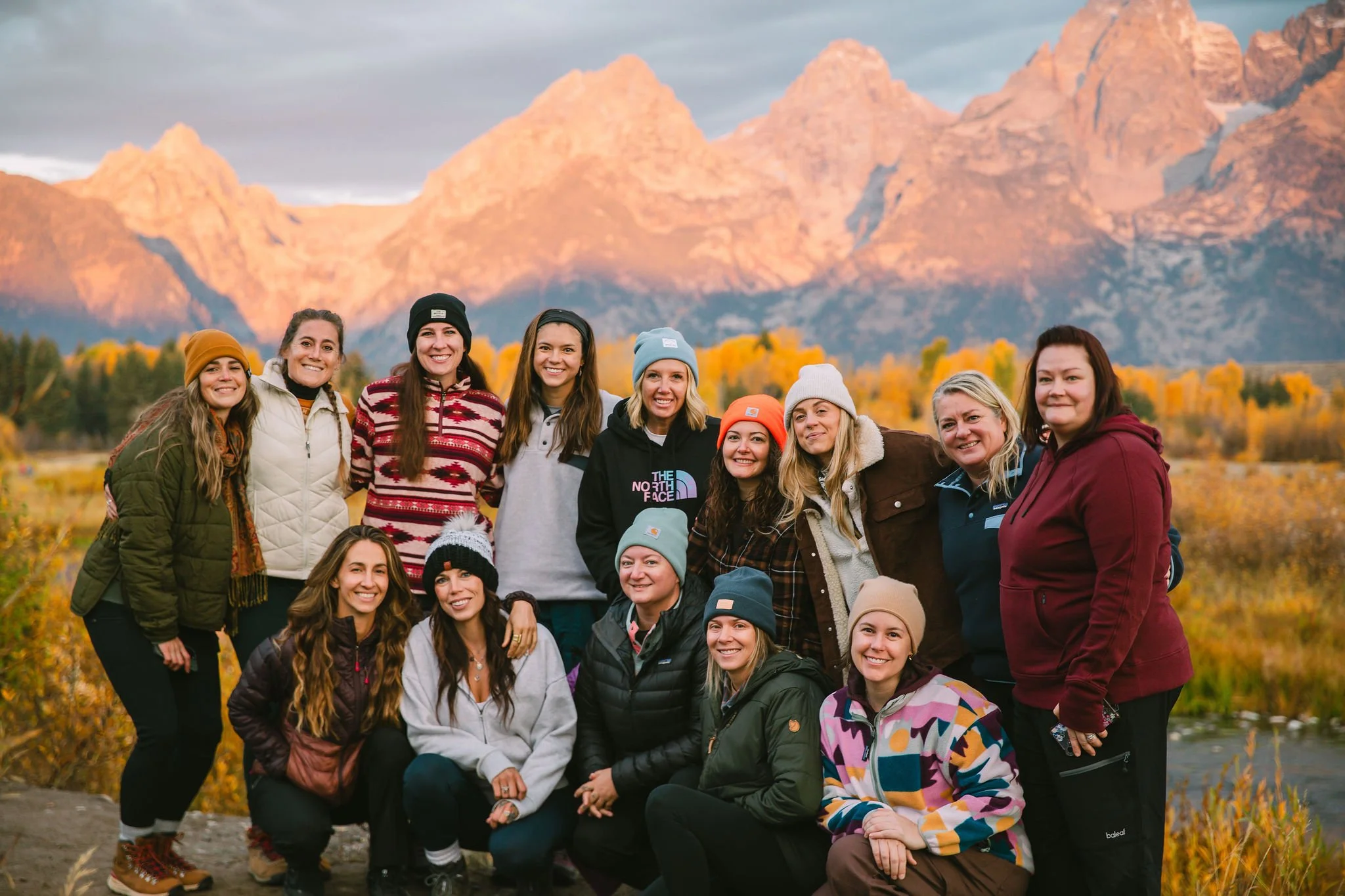 A group of ten women hiking in a mountainous landscape with green hills, trees, and rocky peaks in the background.