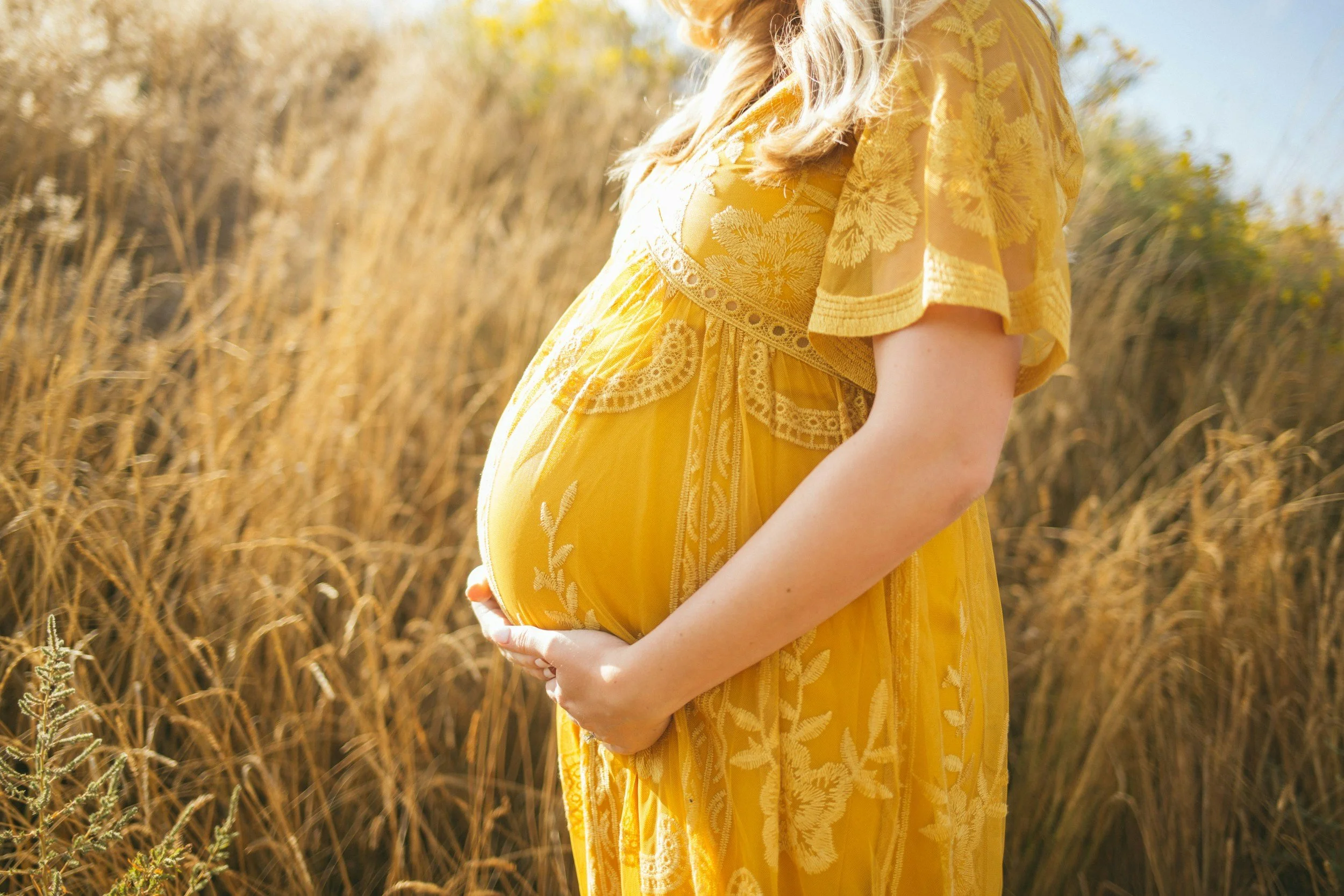 pregnant woman in yellow dress holding belly and standing in field of wheat