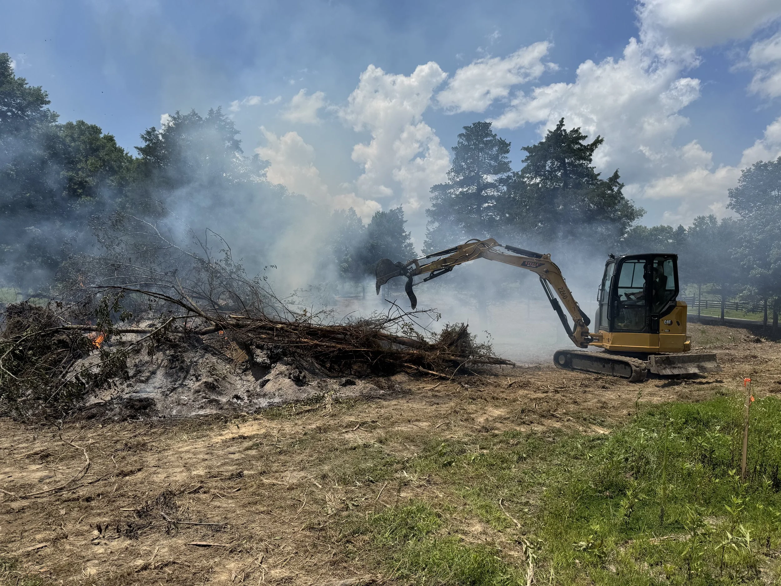 A small yellow excavator removing burnt branches and debris from a smoldering fire site outdoors, with smoke and a partly cloudy sky in the background.
