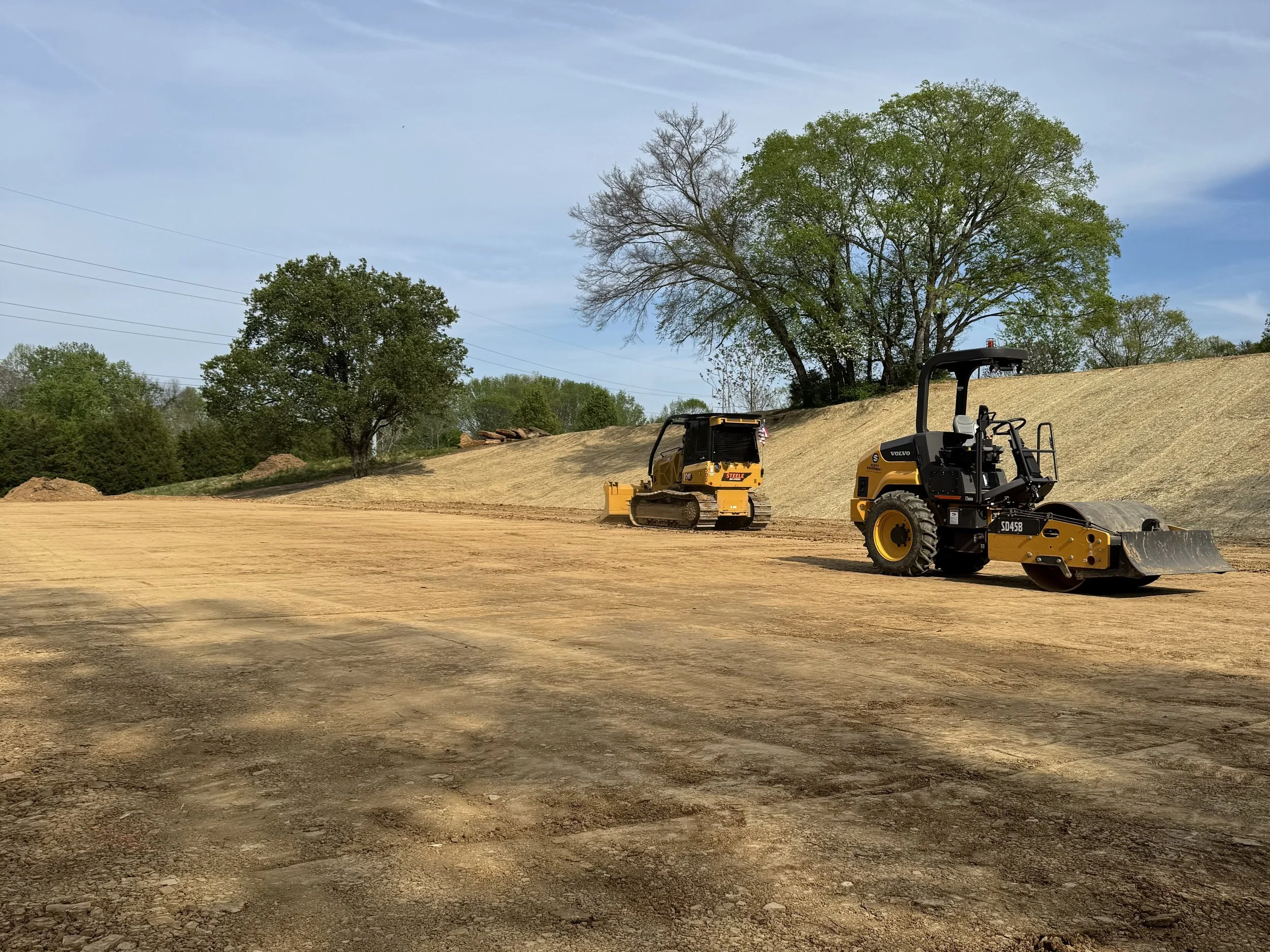 Construction site with two bulldozers working on the dirt ground, large trees in the background, and a partly cloudy sky.