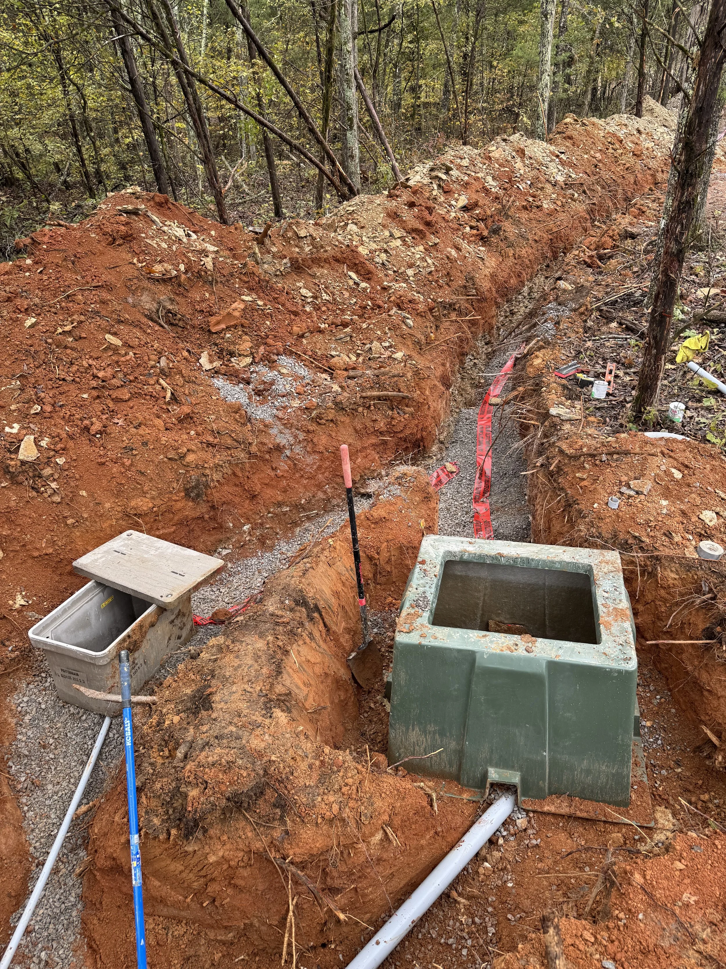 An outdoor construction site in a wooded area with orange dirt, tools, and pipes. A trench is dug, with a white pipe laying inside, and equipment nearby.
