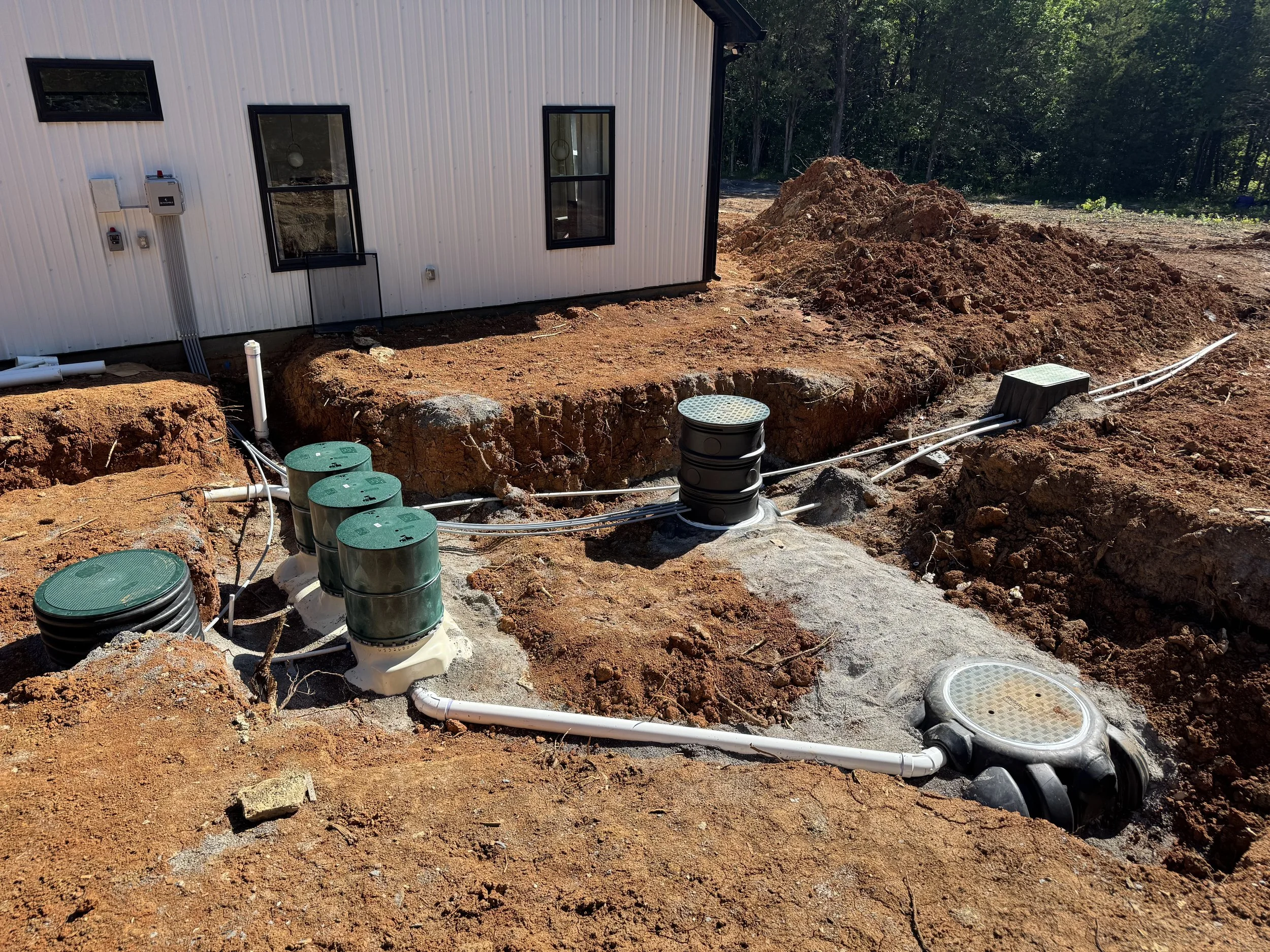 Construction site with dug-up ground, pipes, and utility access points, next to a building with metal siding and black-framed windows.