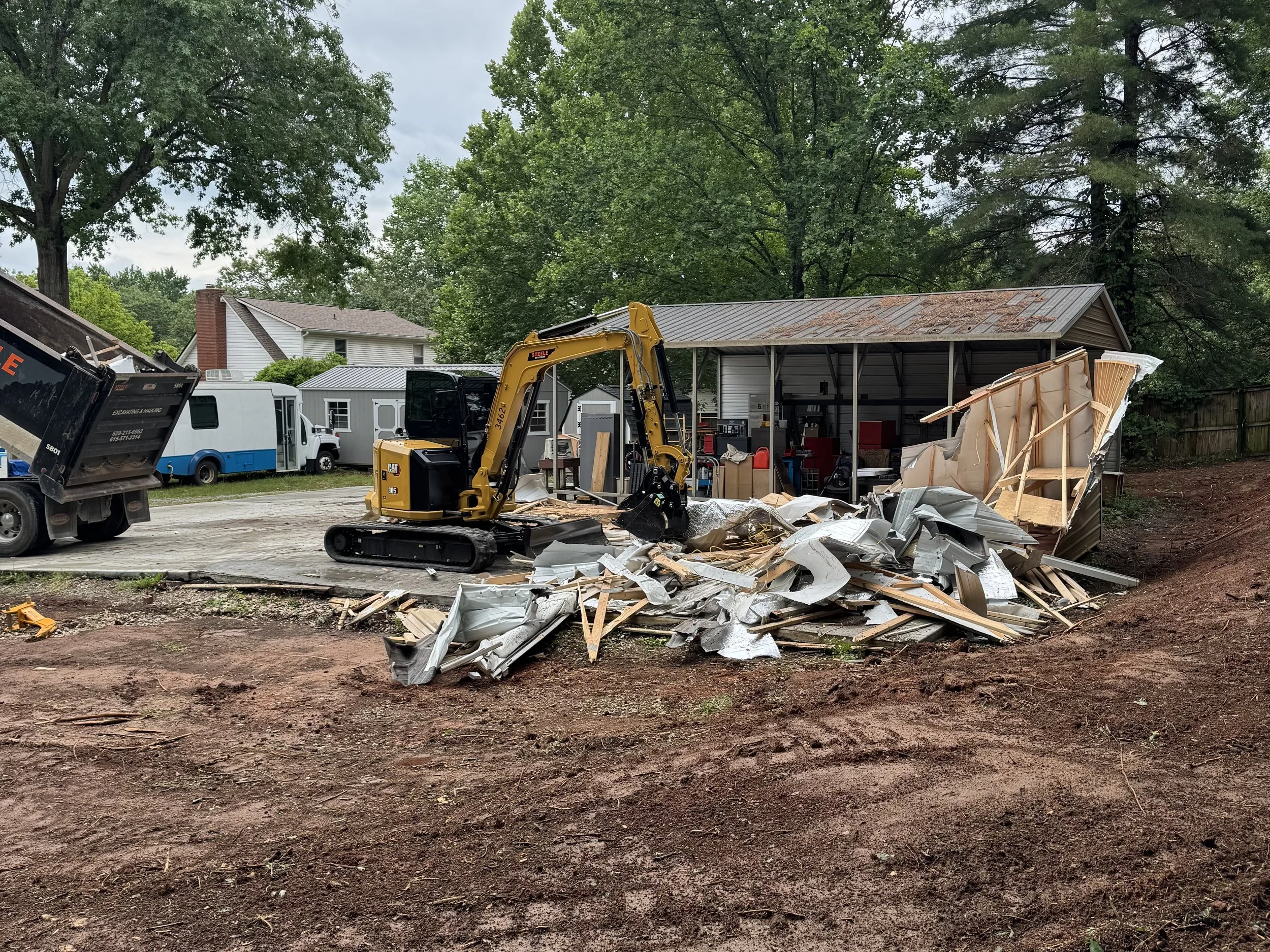 A construction site with debris and broken wood, a yellow mini excavator, and a shed with a rusty roof surrounded by trees and residential houses.