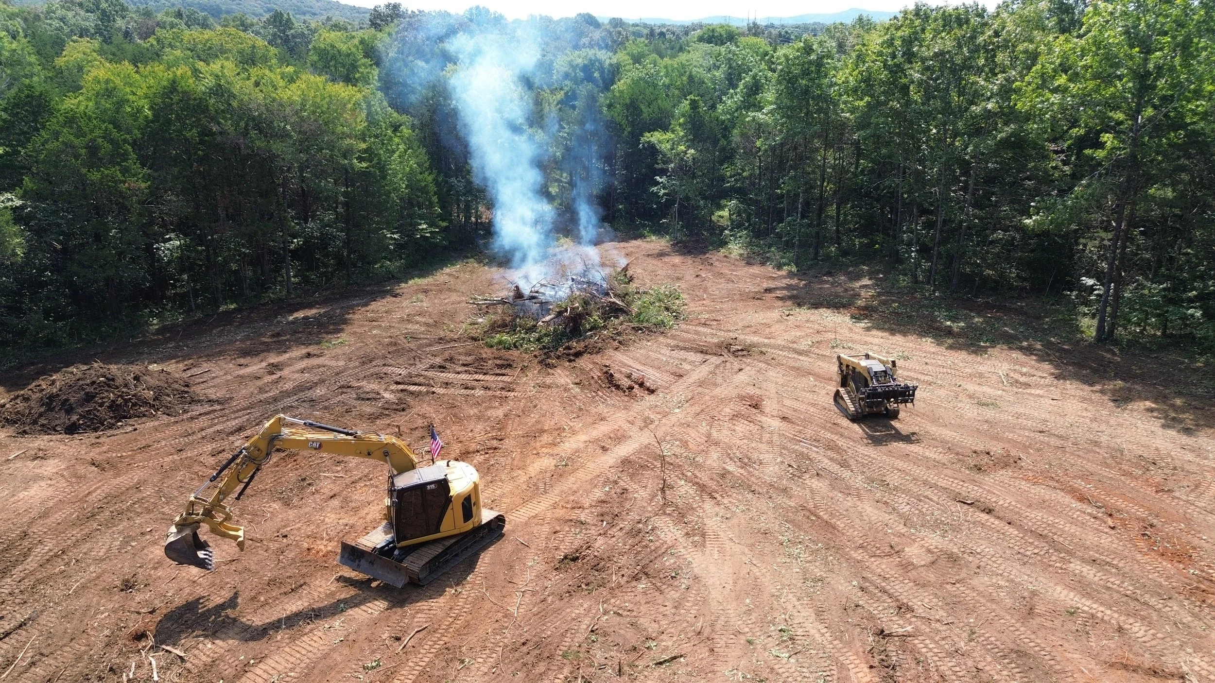 Construction site in a cleared area with two small excavators working near a pile of wood and brush, smoke rising from the fire, surrounded by a dense forest in the background.