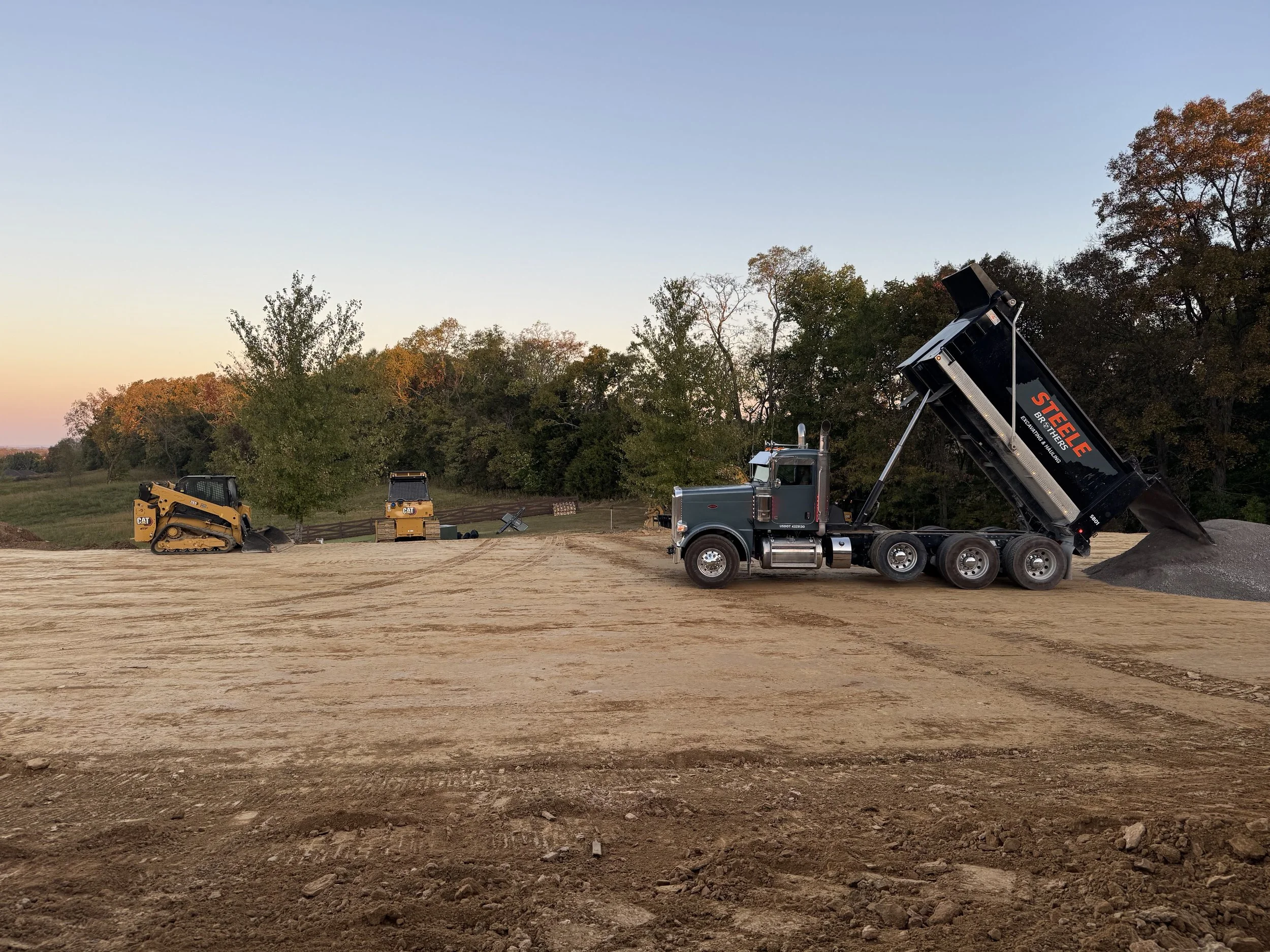Construction site with a bulldozer, a dump truck, and other machinery, surrounded by trees with sunset lighting.