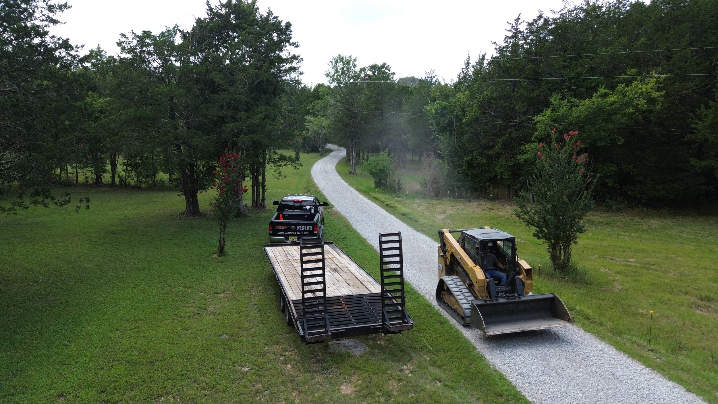 A gravel driveway with a pickup truck, and a construction loader on a grassy area surrounded by trees.