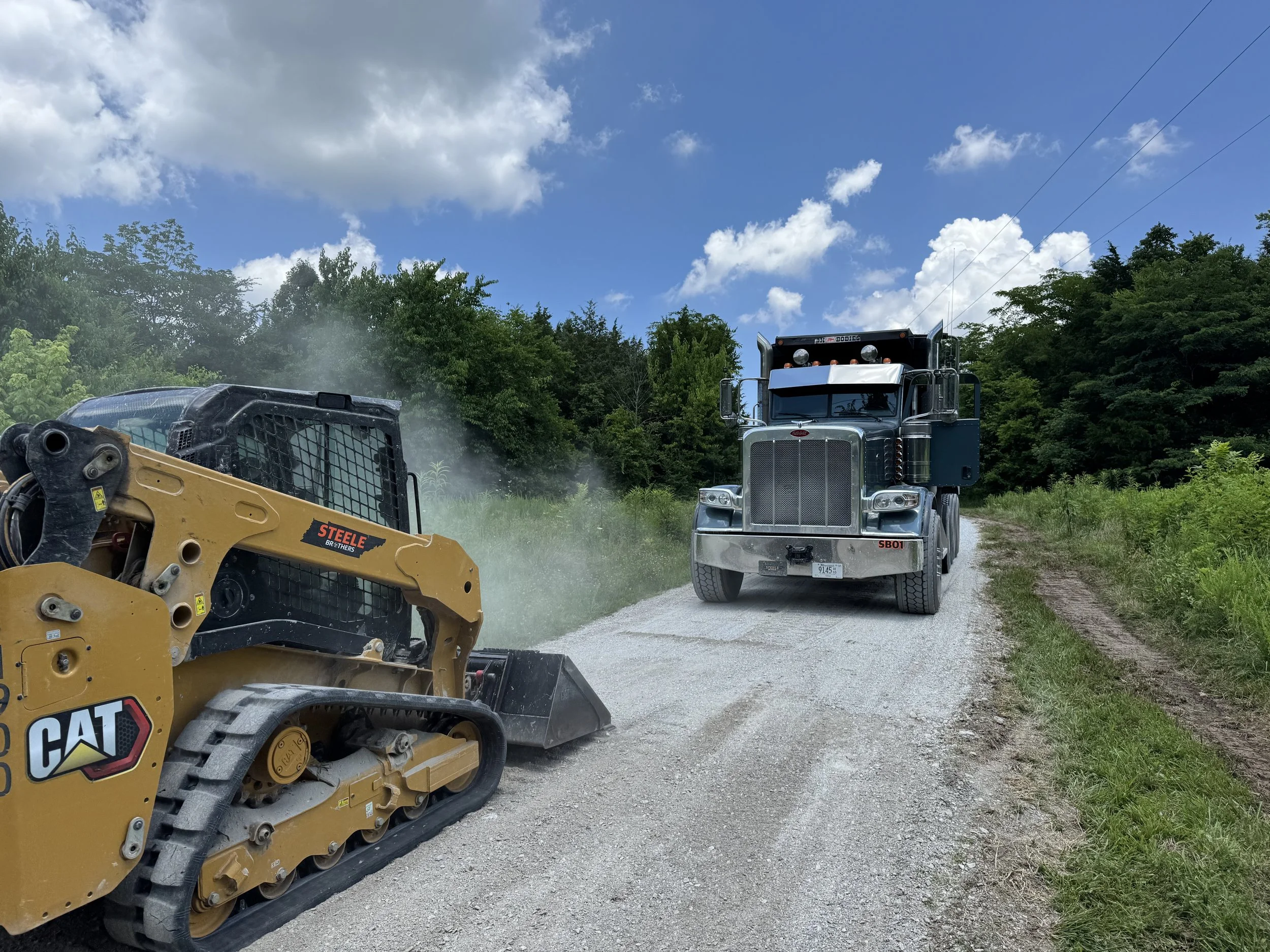 A large black semi-truck is stopped on a gravel rural road, facing a yellow Caterpillar compact track loader that is operating on the side of the road, kicking up dust. The background shows green trees and a partly cloudy blue sky.