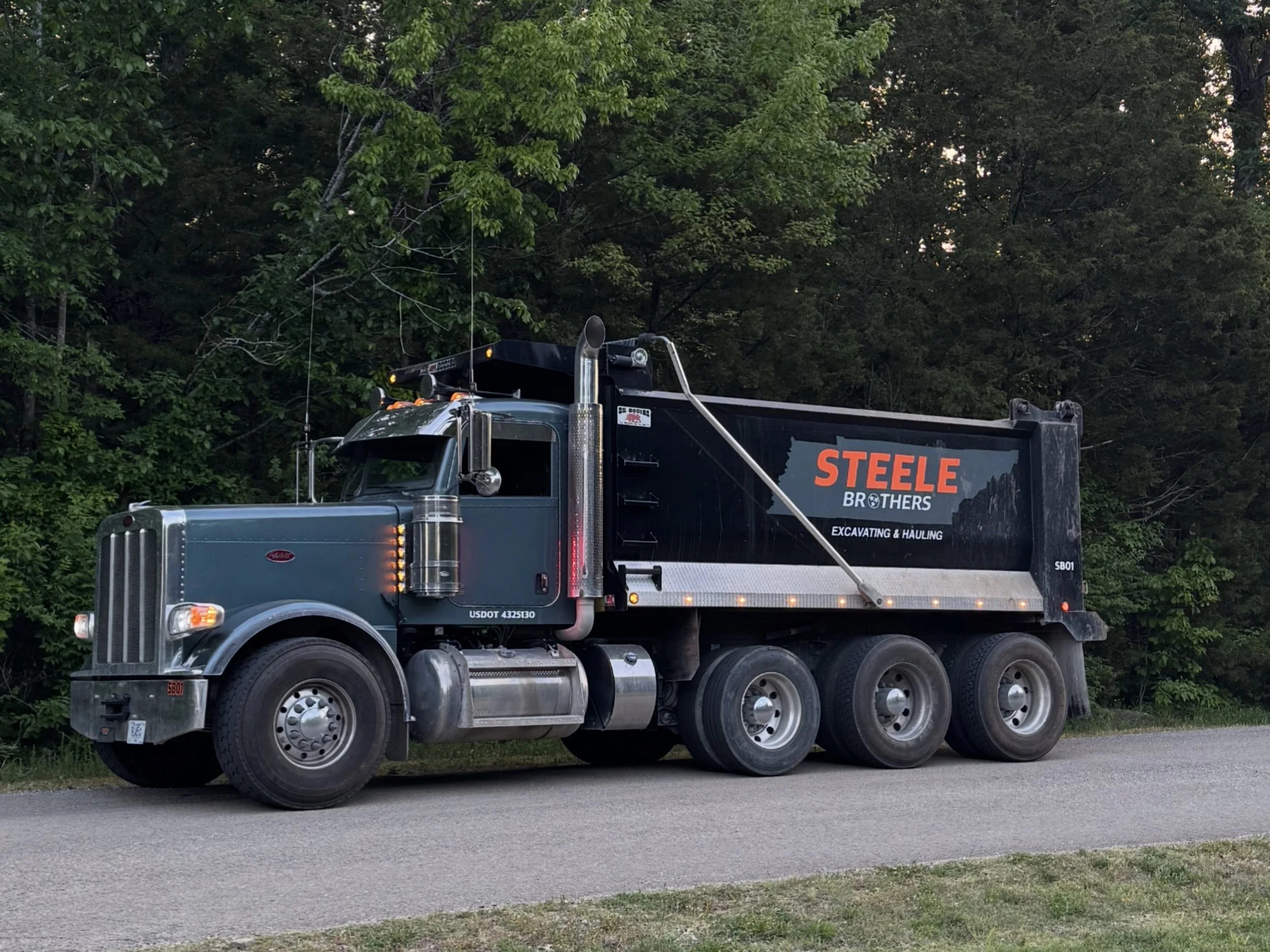 A large black truck with a flatbed and workers' company branding, parked on a paved road beside grass and dense green trees.